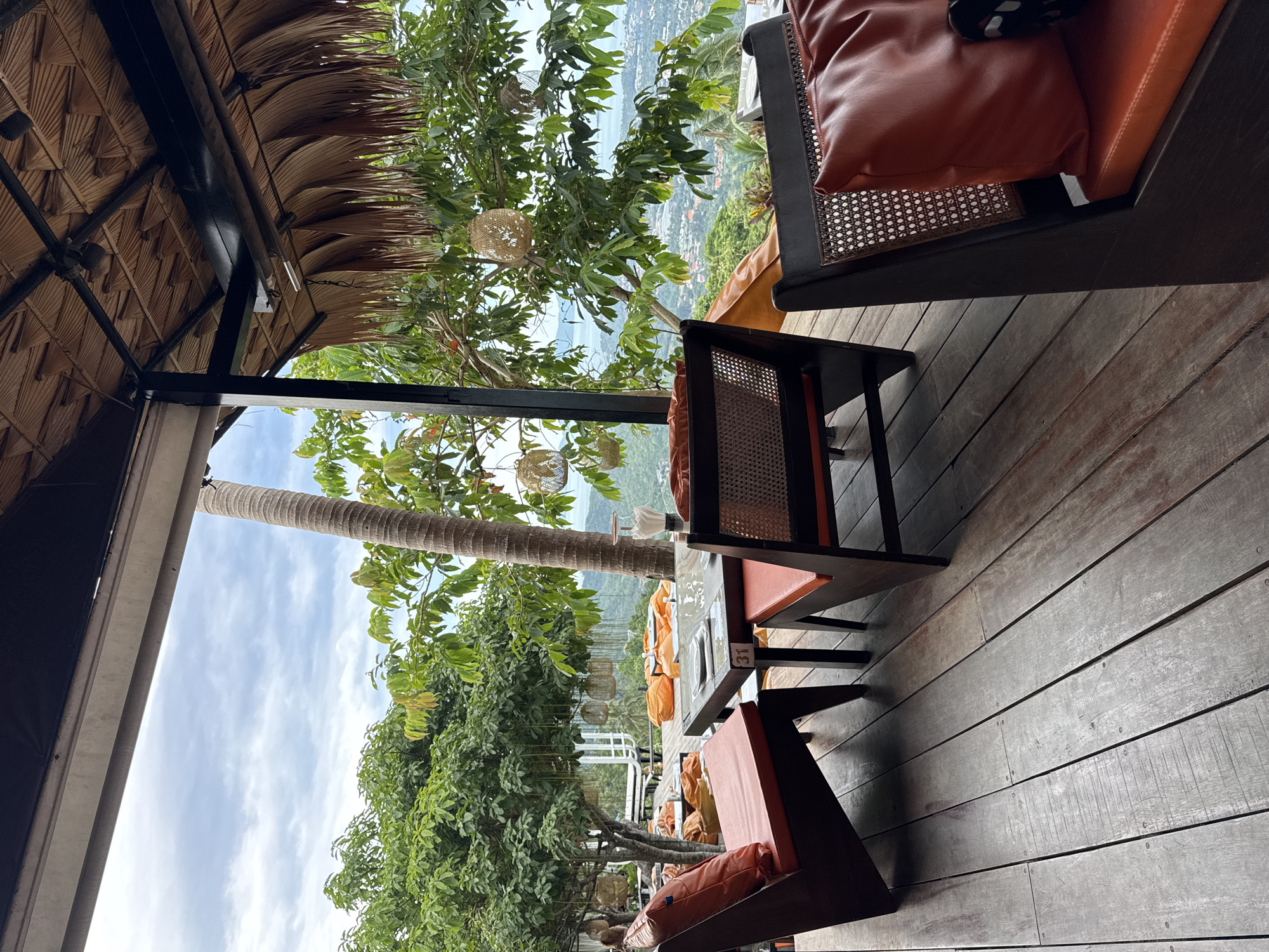 An outdoor seating area with a thatched roof overlooks a tropical landscape and city. The area is furnished with wooden chairs and tables, some with orange cushions. The chairs have woven rattan backs and arms. There are also orange floor cushions visible in the background. Large trees with dense green foliage surround the seating area. Several woven lanterns are suspended from the trees. The ground is made of weathered wooden planks. The sky is cloudy.
