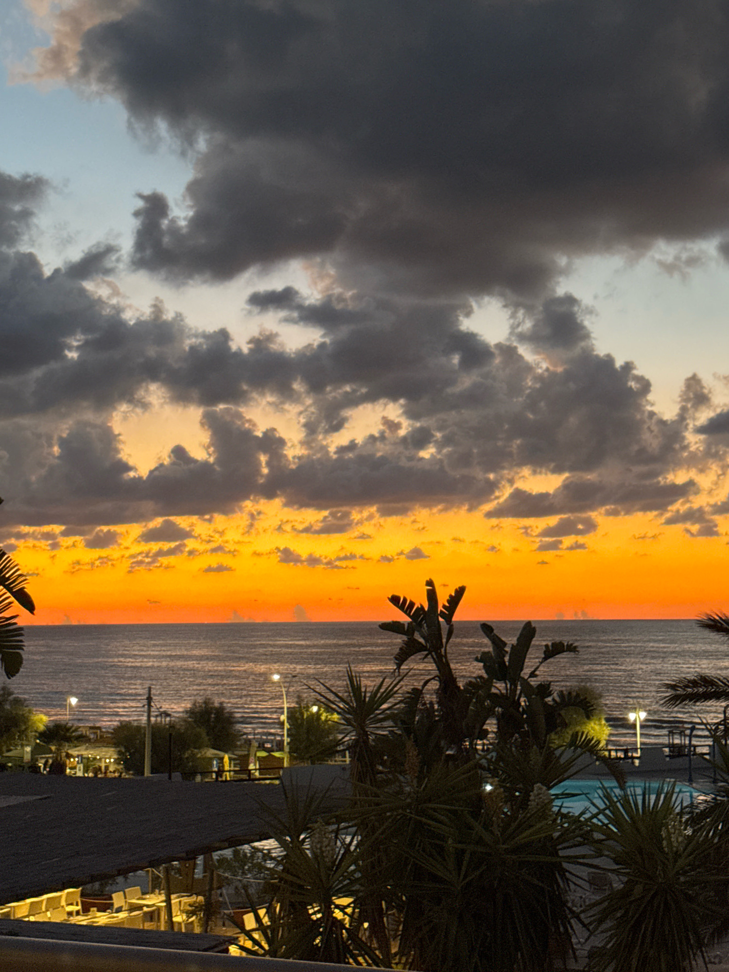 An evening sky transitions from a bright orange glow near the horizon to dark, heavy clouds overhead. The sea stretches out before the viewer, its surface rippled by gentle waves. In the foreground, lush palm trees and other tropical vegetation obscure parts of the view. Beyond the foliage, a resort or beachfront area is visible, with buildings, outdoor seating arrangements, and illuminated street lamps. The atmosphere suggests a peaceful, tropical evening on the coast of Isola delle Femmine, Italy.