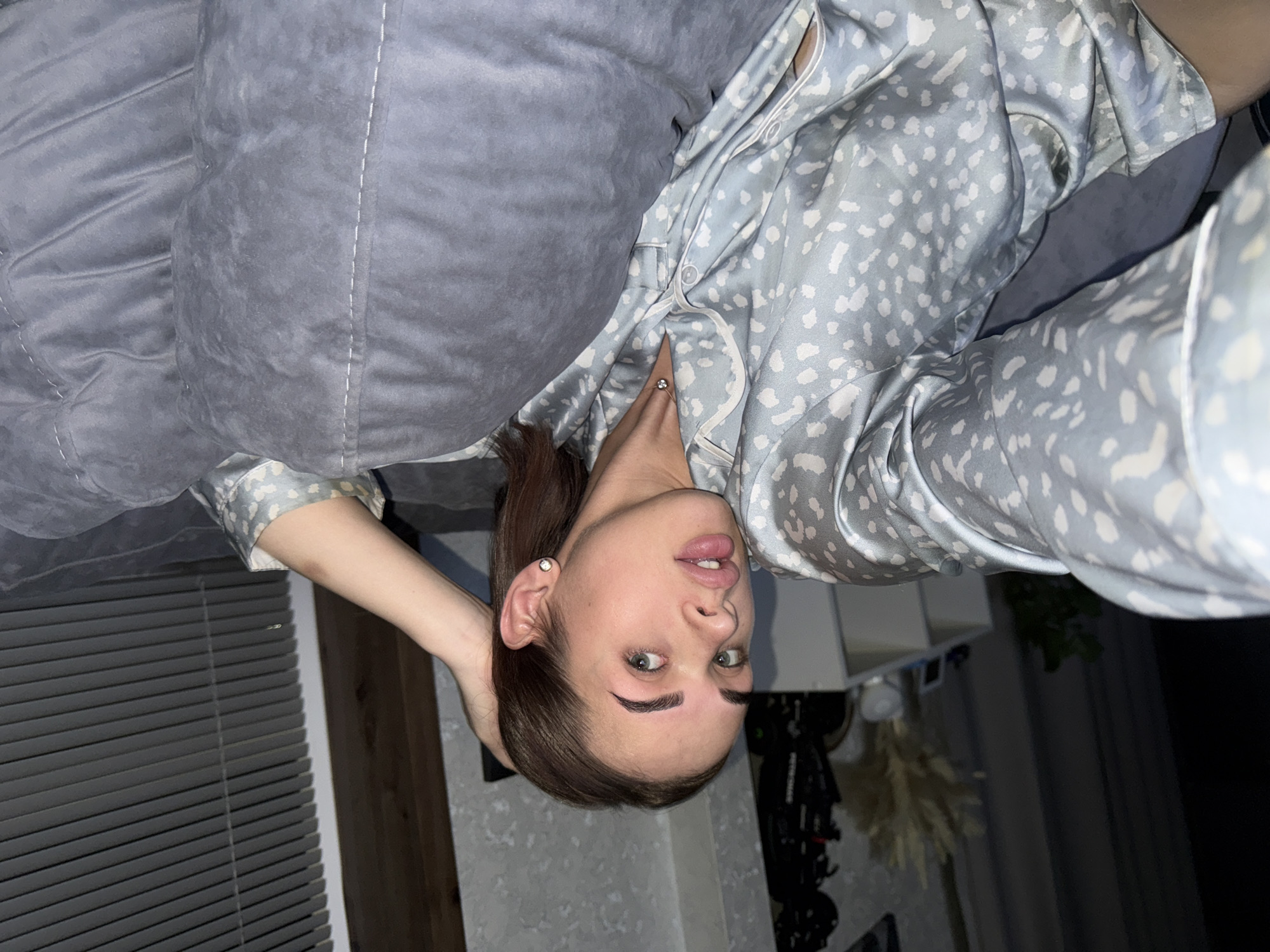 A young woman with brown hair and green eyes is lying on a gray, quilted cushion or pillow while wearing a blue and white spotted pajama top. Her head is tilted back, and she is looking directly at the camera. Her right hand is behind her head. A window with closed horizontal blinds is visible on the left side of the frame. Behind the woman, there is a white wall and dark furniture or decor. This image was taken in Лобаново, Russia.