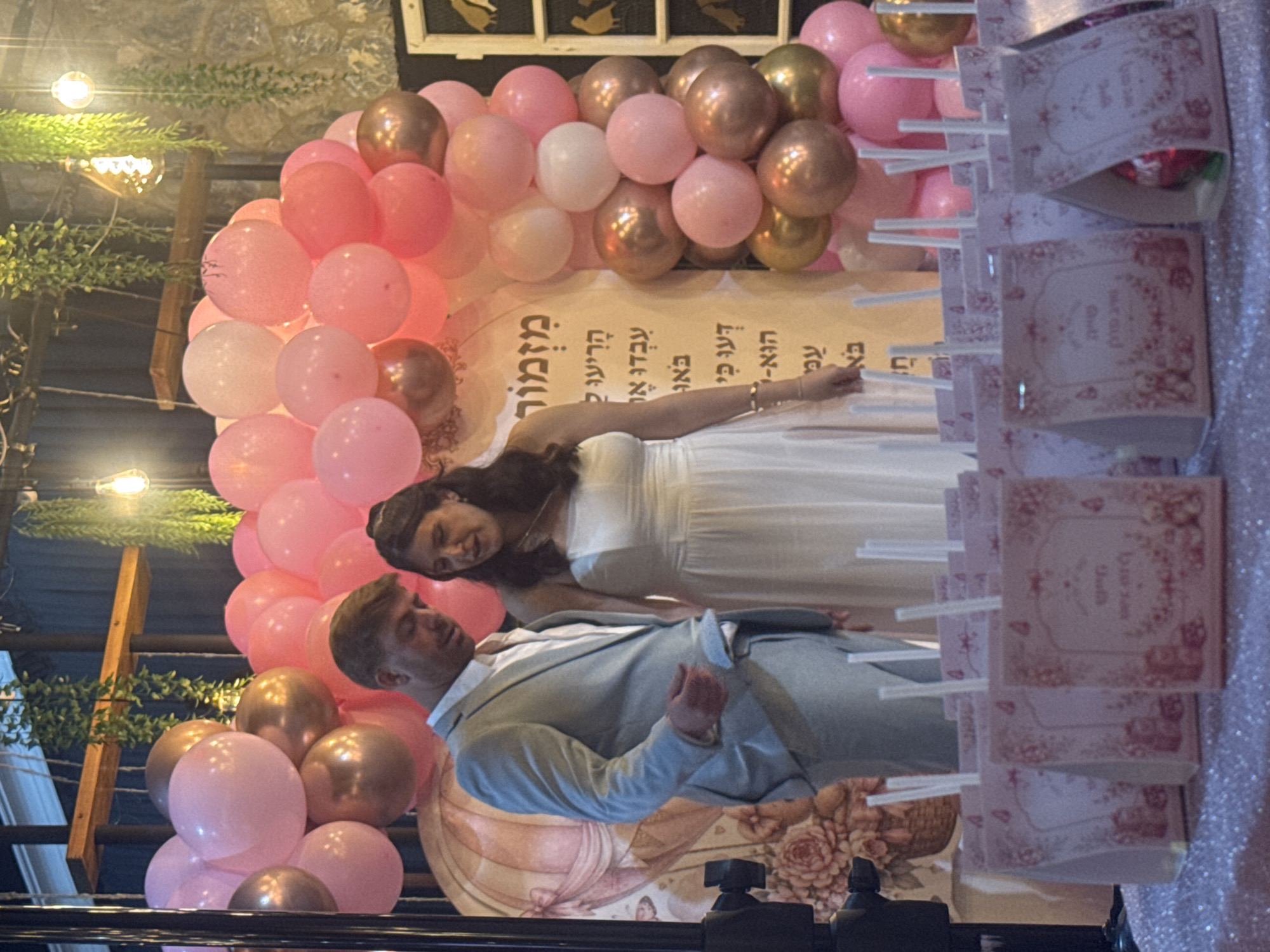 In Migdal HaEmek, Israel, a man in a light gray suit and a woman in a white dress stand in front of a balloon arch. The balloons are pink and gold. Behind them is a backdrop featuring a hot air balloon design and Hebrew text. In the foreground, several pink gift bags with straws are arranged on a table. The event appears to be an outdoor celebration.