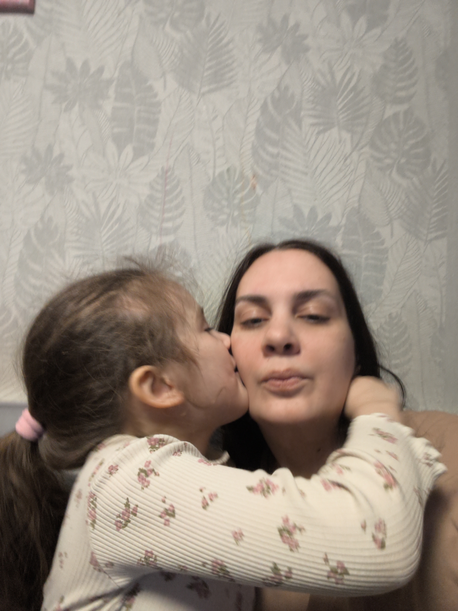 A young girl with brown hair pulled back in a ponytail kisses a woman's cheek. The woman has dark hair and fair skin. Both are indoors, with a light-colored wallpaper featuring a subtle leaf pattern in the background. The child is wearing a white, long-sleeved shirt with a floral print. The woman is wearing a light brown top. Location is indiscernible in this photo and information of whether this photograph was taken in Chaikovsky, Russia is unavailable.
