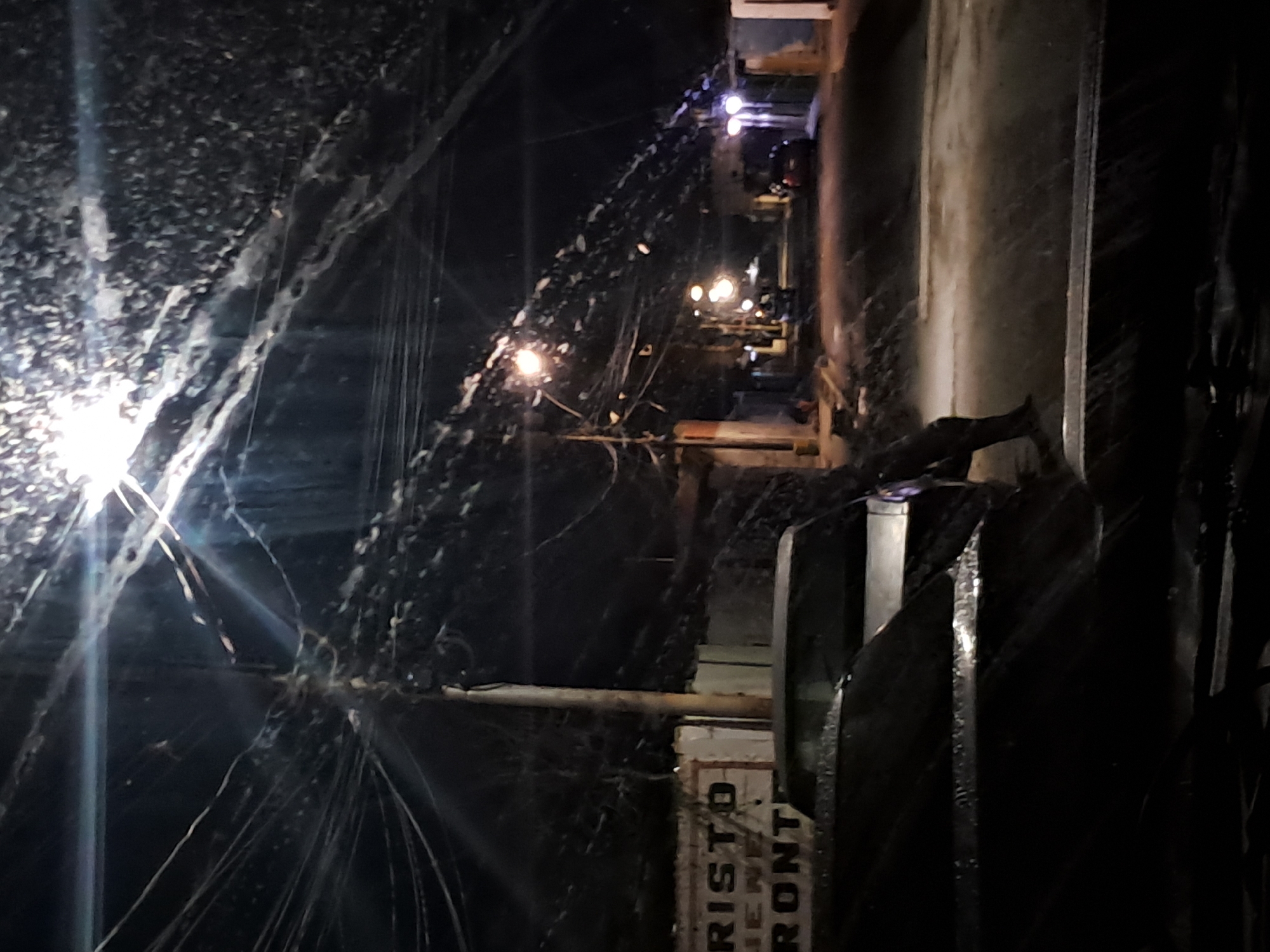This image captures a nighttime street scene in Cumana, Venezuela, likely during or after a heavy rain. The foreground is dominated by the dark, reflective surface of what appears to be the hood of a vehicle, with streaks of water visible, suggesting recent precipitation. In the background, a street is illuminated by several streetlights, casting a glow on the wet pavement and creating reflections. Several buildings line the street, some with signs that are partially visible. One sign appears to read "CRISTO VIENE PRONTO". A utility pole with a dense network of wires stretches across the frame. A few cars are parked or driving on the street, their headlights and taillights visible. The overall atmosphere is dimly lit and damp, with a sense of urban quietude.