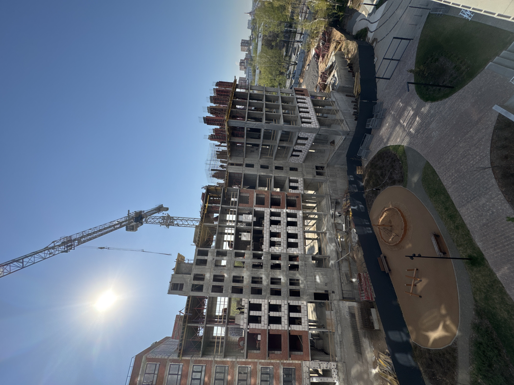 The image shows an elevated view of a construction site in Izhevsk, Russia. A partially constructed high-rise building dominates the scene, featuring a concrete skeleton with some brick and block infill. A large construction crane is positioned adjacent to the building. Ground-level construction materials and equipment are visible. A newly constructed park area featuring a playground, benches, and walkways is directly adjacent to the construction site. Beyond the immediate construction zone, glimpses of other buildings and trees indicate a mixed urban landscape under a clear blue sky with a bright sun.