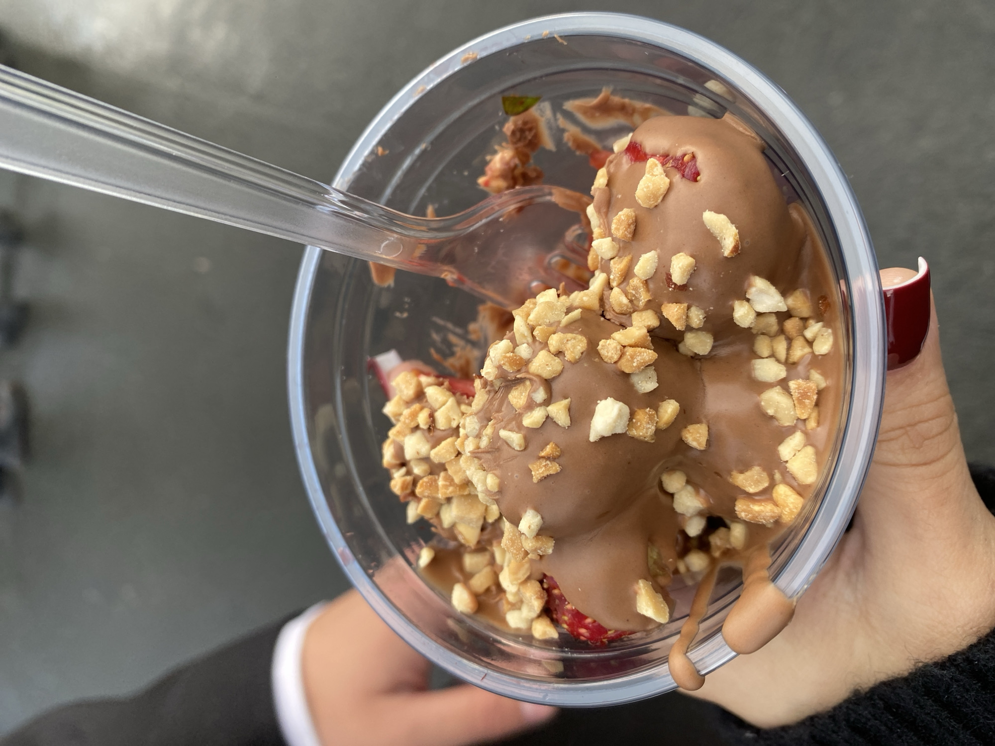 The image shows a close-up, overhead view of a clear plastic cup filled with a chocolate-covered dessert, likely strawberries, topped with chopped nuts. A clear plastic fork is partially submerged in the dessert. The dessert appears to be melting slightly, with chocolate dripping down the side of the cup. The cup is held by a person whose hand, with dark red nail polish, is visible on the right side of the frame. The background is out of focus and appears to be outdoors, possibly on a paved surface, suggesting a casual setting. The lighting indicates it is daytime, but specific weather conditions cannot be determined. There is no visible text in the image. The overall impression is of a sweet treat being enjoyed outdoors.