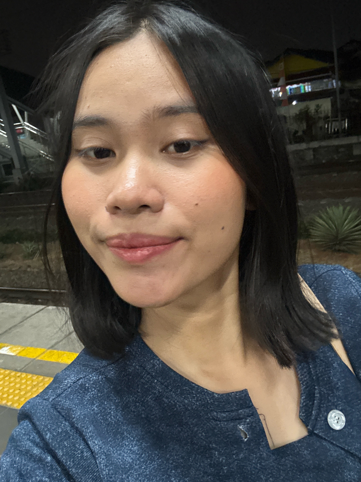A close-up selfie shows a young Indonesian woman with shoulder-length dark hair and fair skin. She is wearing a dark blue textured top. Her makeup includes blush and lip gloss. In the background, out of focus, there are train tracks and a building with lights. The scene appears to be at night, possibly at a train station. The presence of train tracks suggests a location in East Jakarta, Indonesia, which has several train stations.