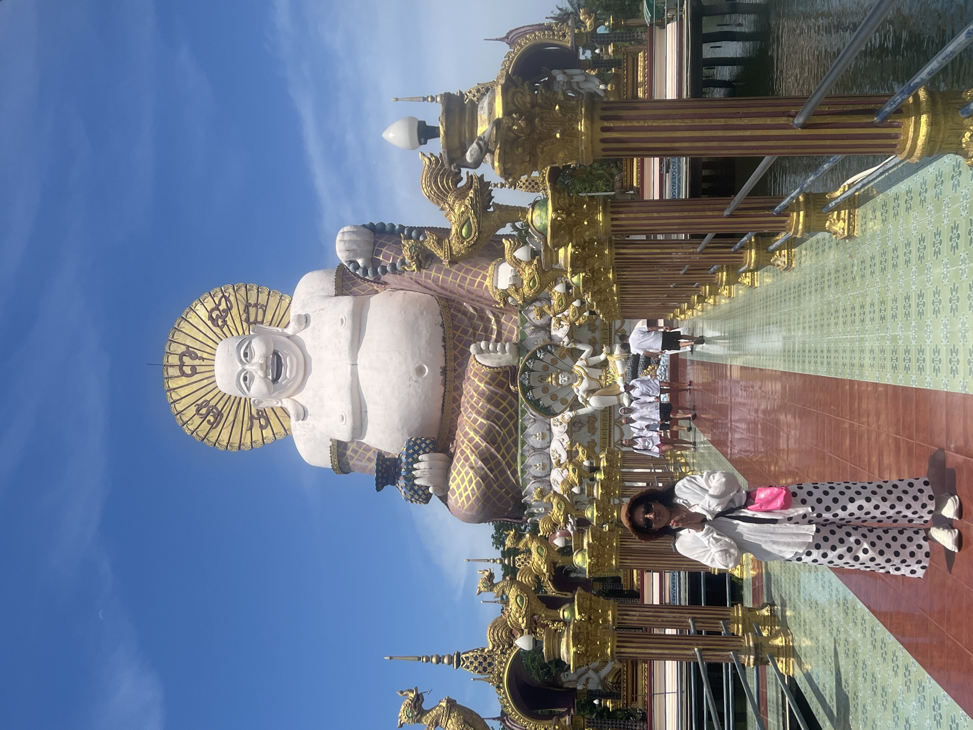 In Ko Samui District, Thailand, a woman stands on a walkway in front of a large statue of a smiling Buddha. The walkway is lined with ornate golden pillars and decorative statues. The woman is wearing a white shirt, polka dot pants, a hat, and sunglasses, and is holding her hands together in a prayer-like gesture. Behind her, several people are walking away from the camera. The statue of the Buddha is seated and adorned with gold and purple fabric, and has a golden halo behind its head. The sky is blue with some clouds.