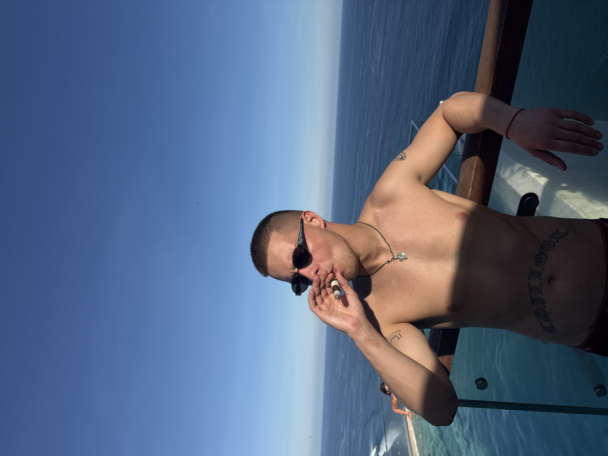A shirtless man with short, cropped hair is pictured on a sunny day, leaning against a wooden railing with a clear glass partition below. He is wearing sunglasses and has a tattoo on his stomach that says "CHILLOUT" and a necklace with a cross. He is holding what appears to be a cigar in his hand. The background shows a horizon line between a light blue sea and a slightly lighter blue sky. The location is Kuta Selatan, Indonesia.