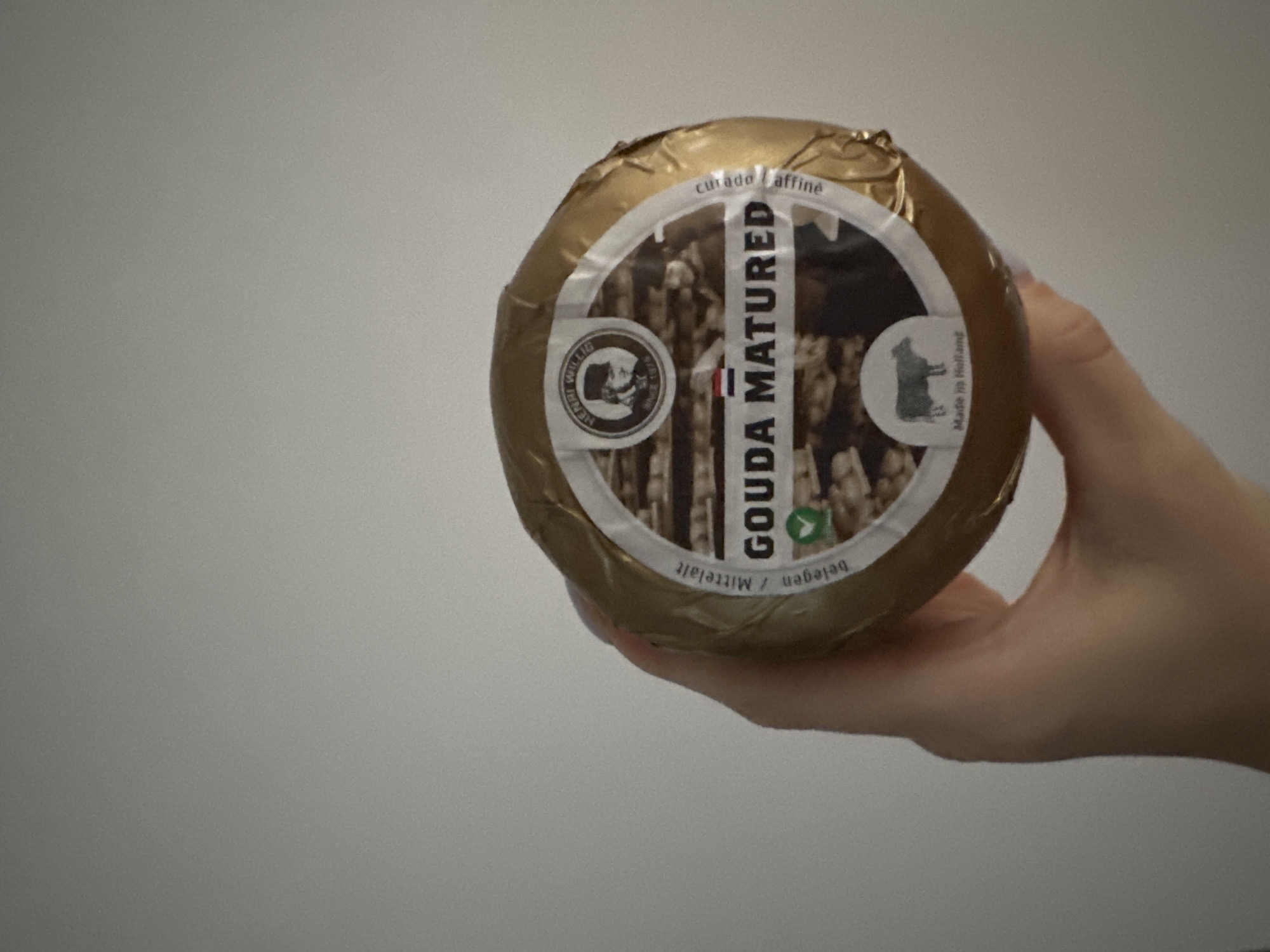 A hand is holding a round wheel of Gouda cheese. The cheese is wrapped in gold foil and has a white label with Dutch text and a picture of a man in a hat. The text on the label reads "GOUDA MATURED." The cheese is a light brown color. The background is a plain white wall. There is no visible indication that this image was taken in Murcia, Spain.