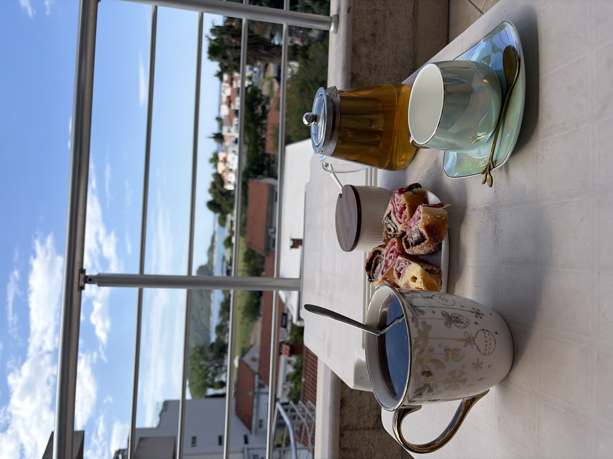 The image depicts a table on a balcony in Budva, Montenegro, laden with tea and pastries. A glass teapot with golden liquid sits beside a pastel blue cup and saucer with a gilded spoon. A small plate of sliced poppy seed rolls is next to a sugar bowl with a wooden lid. A white mug with gold accents, filled with a dark liquid and a spoon, is also on the table. The balcony railing frames a view of Budva, revealing rooftops, trees, and the coastline. The sky is blue with scattered clouds.