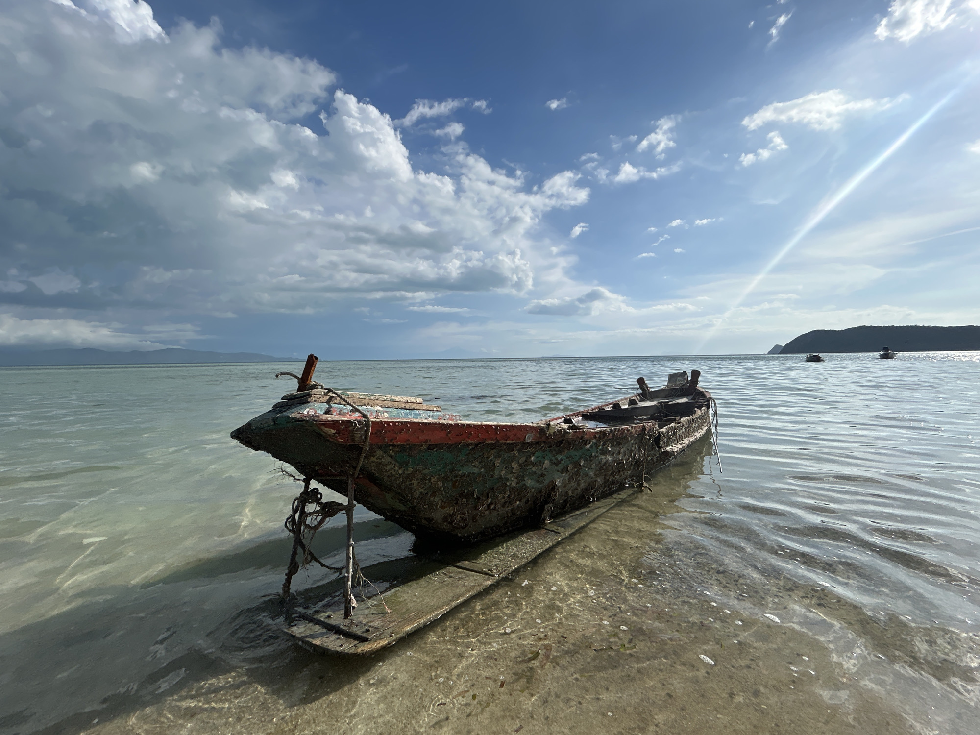 A deteriorated wooden boat rests in the shallow water along the shoreline of ตำบลเกาะพะงัน, Thailand. The hull is covered in marine growth with remnants of faded red paint. A crude wooden platform supports the boat. In the background, the sea stretches to the horizon under a sky with scattered cumulus clouds and bright sunlight. Several distant islands and small boats are visible. The water is clear and shallow near the shore, revealing the sandy seabed.