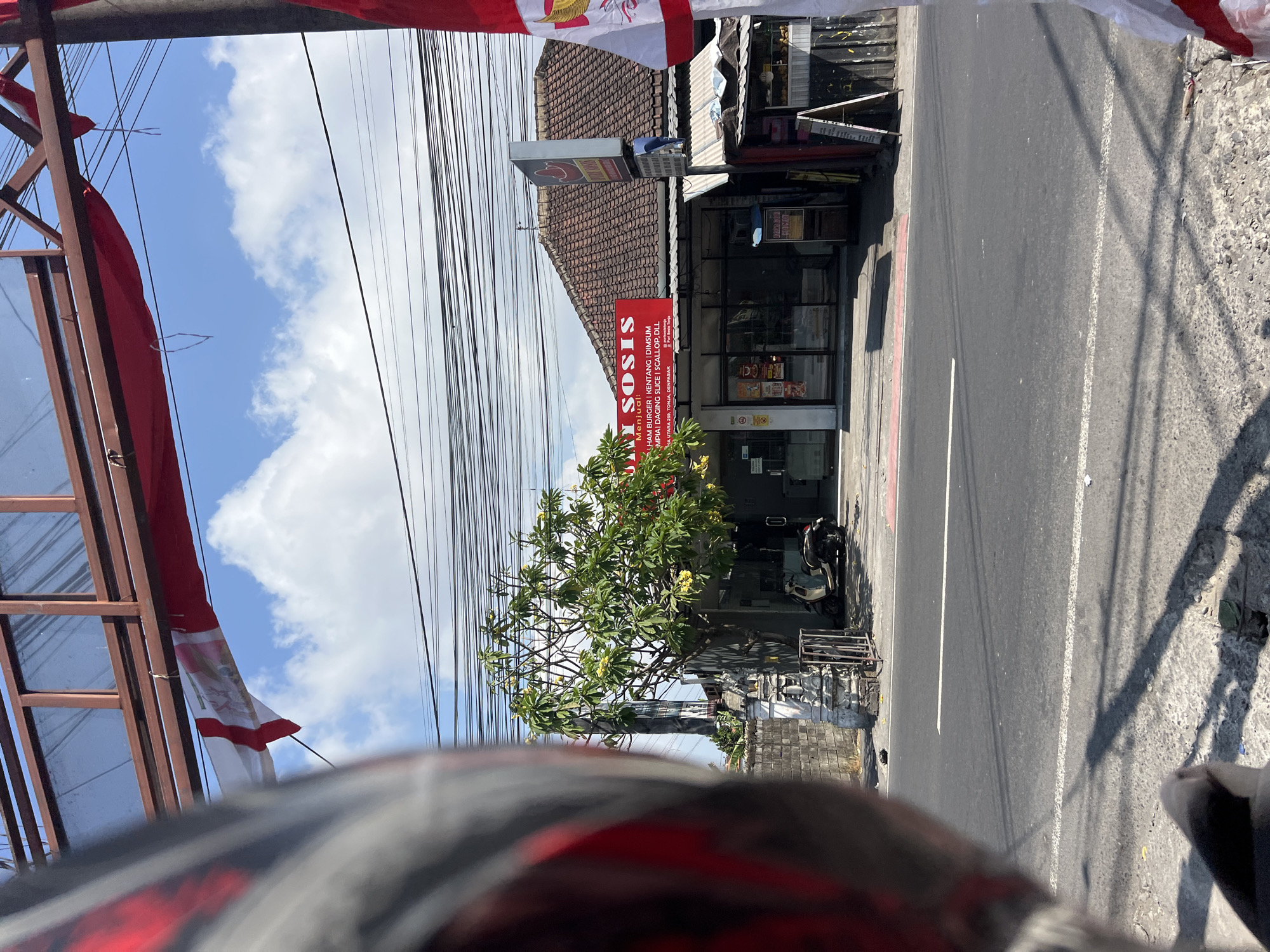 The image captures a street scene in Denpasar, Indonesia, on a sunny day with scattered clouds. In the foreground, the left side of a person's helmet, with red and grey detailing, is visible, suggesting the viewer is on a motorcycle. Across the asphalt road, a single-story building with a tiled roof houses a business displaying a red sign with white text that reads "Y SOSIS". Below this, smaller text indicates "MENJUAL" and lists items like "HAM BURGER | KENTANG | DIMSUM | UMPAI | DAGING SLICE | SCALLOP, DLL". The building has large glass windows and doors, with a scooter parked in front of it. Adjacent to the building is a small stall or kiosk with a white roof and a display board. A frangipani tree with green leaves and yellow flowers stands to the left of the business. Overhead, a tangle of black electrical wires crisscrosses the sky. Two red and white flags, likely Indonesian flags, are visible, one hanging from a structure above the business and another partially obscuring the right side of the frame. A stone wall with decorative carvings is situated to the left of the street.