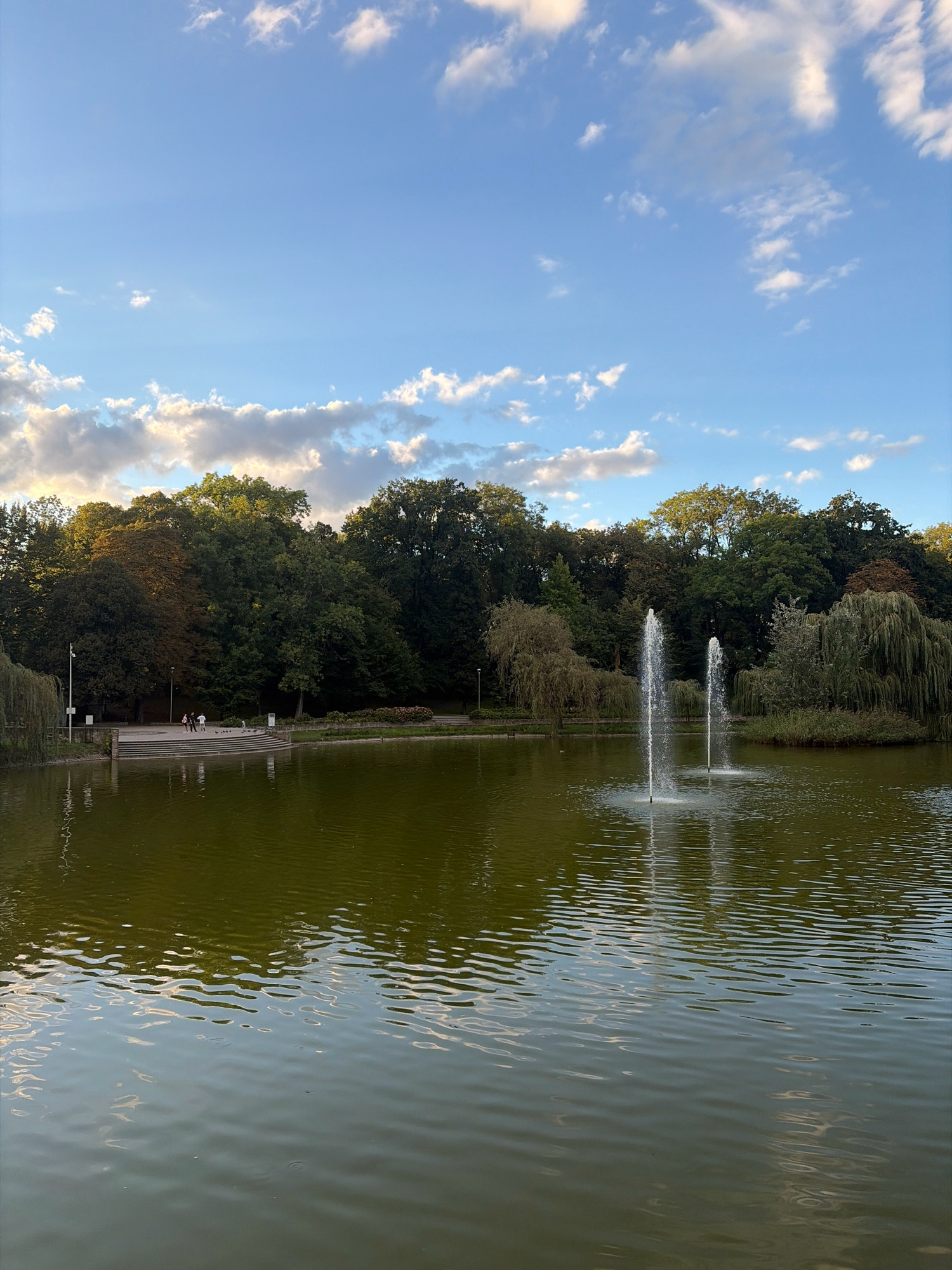 The image depicts a serene park scene with a large pond and lush green trees lining its banks. Two fountains are actively spraying water into the pond, creating a dynamic visual element. On the left side of the pond, a paved walkway and steps lead down to the water's edge, where a few people are seen walking and interacting. The sky above is a clear blue, dotted with scattered white clouds, suggesting pleasant weather. The overall atmosphere is tranquil and inviting, characteristic of a public park.
This image was taken in Racibórz, Poland, specifically at the OSiR Racibórz park.