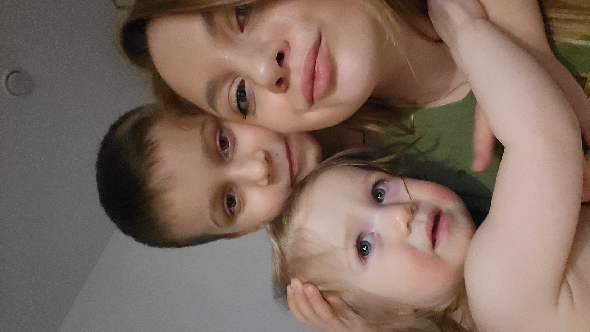 The image depicts a selfie of a woman and two children. The woman appears to be fair-skinned with blonde hair and is positioned at the top of the frame. A young boy with short, dark hair is nestled beside her. A baby with blonde hair is at the bottom of the frame. The background is a light gray wall with a circular fixture on it. There is no contextual information in the image to suggest a location in the city of Chaikovsky, Russia.