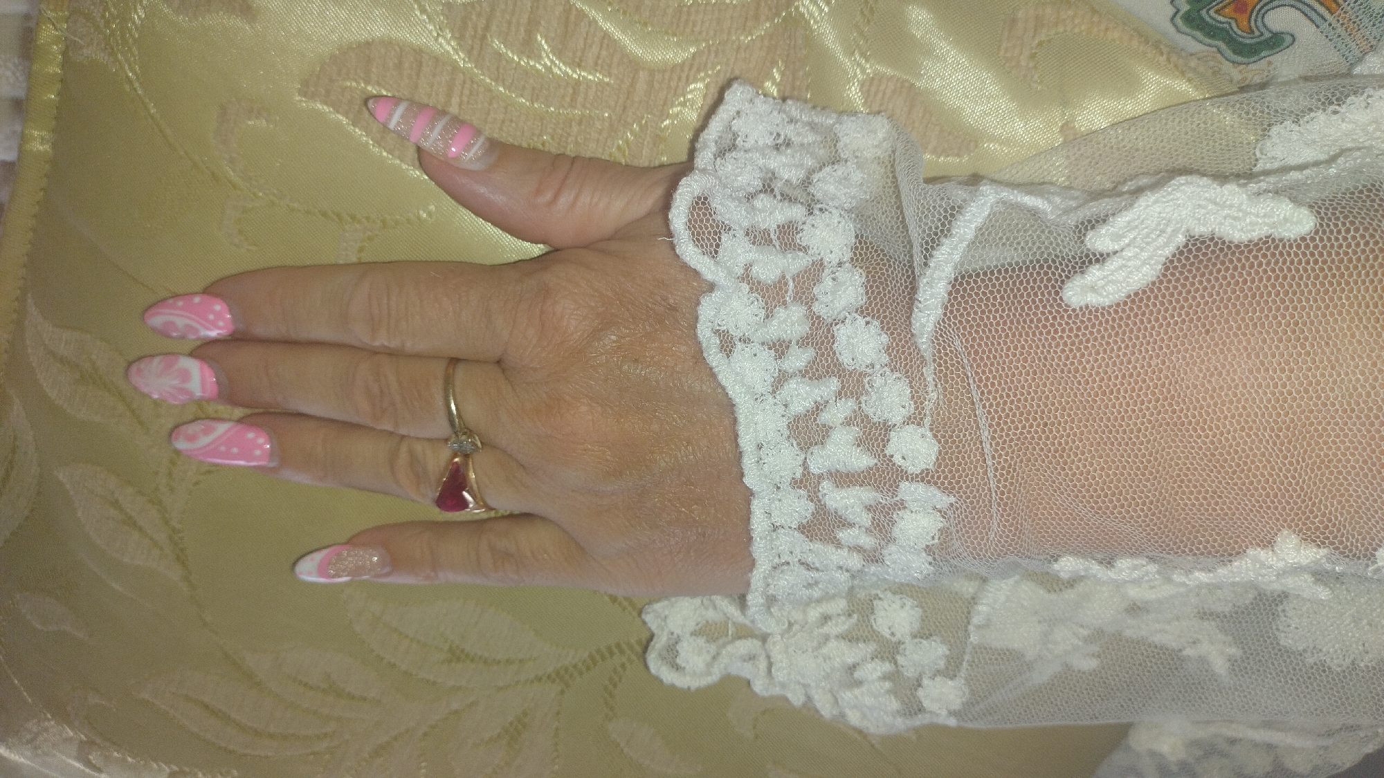 The image shows a close-up view of a person's left hand adorned with decorated fingernails and jewelry, positioned against a textured, light-colored fabric. The fingernails are painted with a pink and white design, featuring stripes on the index finger and floral or abstract patterns on the other visible nails. A ring with a red teardrop-shaped stone and smaller clear stones is worn on the ring finger. A section of white lace trim is visible along the wrist and forearm, suggesting it might be part of a garment like a sleeve or glove. The background fabric has a subtle sheen and a patterned texture, possibly silk or satin, in a muted yellow or cream color. The lighting appears to be artificial, and there are no clear indicators of time of day, weather, or specific location, other than the general context of Perea, Greece. The overall impression is one of personal adornment and detail, focusing on the aesthetics of the hand and its accessories.