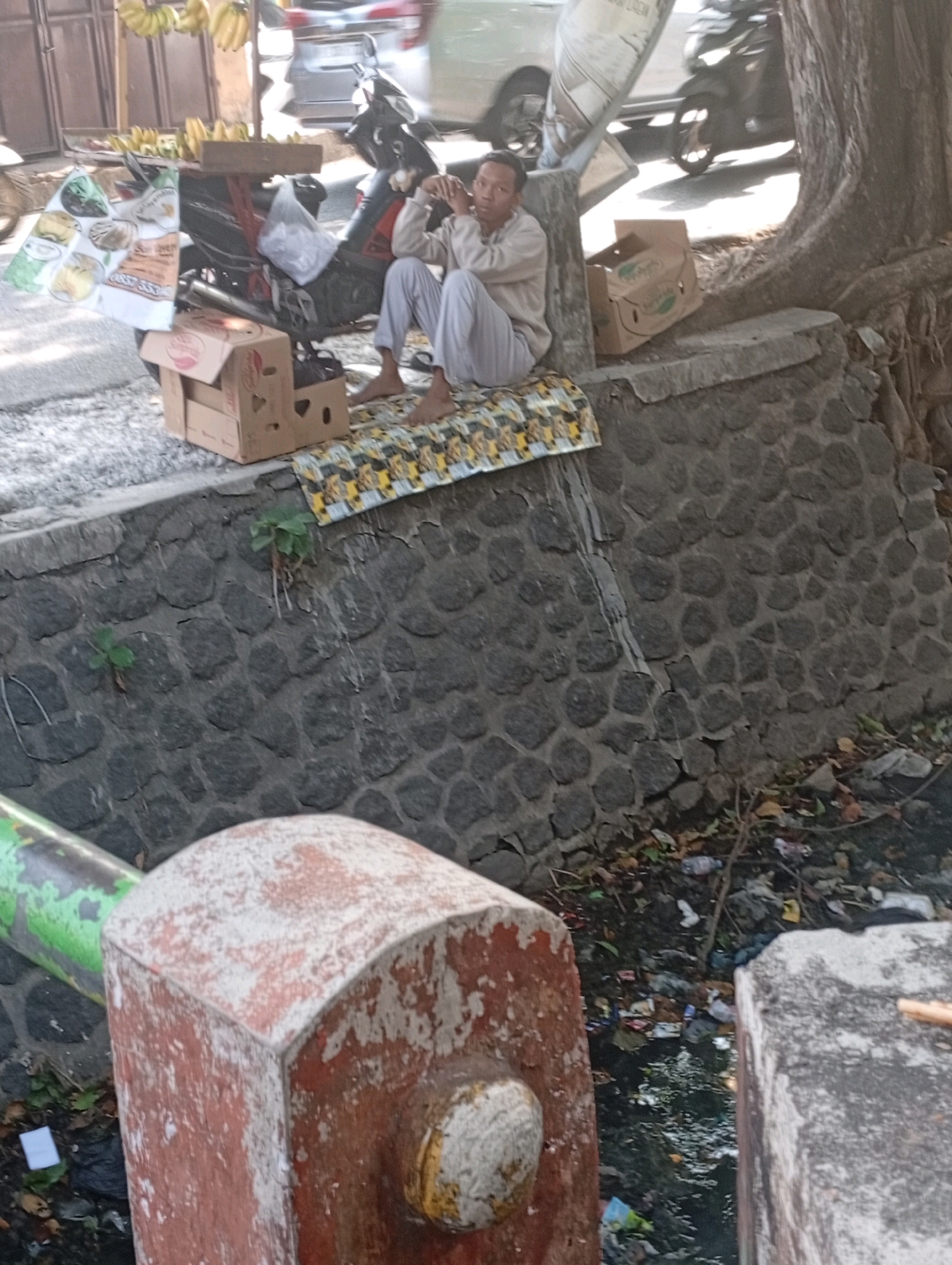In the city of Jogosari, Indonesia, a man sits on a stone wall next to a motorcycle. He is wearing light-colored pants and a long-sleeved shirt, and his legs are crossed. He is looking to his left, with his hands clasped in front of him. A patterned blanket is laid out on the wall where he is sitting, and a cardboard box is placed to his right. The stone wall appears to be part of a drainage ditch, which is filled with trash and debris, including plastic bottles and wrappers. A large tree with exposed roots is behind the man, and a large, weathered red and white structure is in the foreground. In the background, a motorcycle is parked, and a blurry vehicle is visible on the road.
