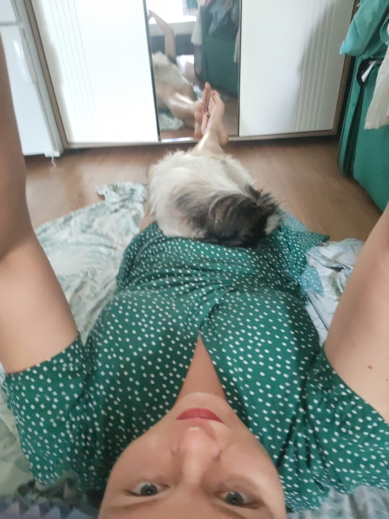 The image shows a first-person perspective of a woman lying on the floor, with a dog resting on her chest. The woman is wearing a green polka-dot dress. In the background, a mirror reflects her legs and feet, as well as part of a closet. The scene appears to be indoors, in a residential setting. There is no visible indication of the city of Izhevsk, Russia, in the image.