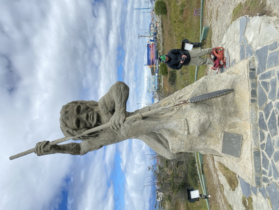 A statue of a native figure holds a spear-like weapon against a backdrop of cloudy skies. A person stands near the statue's base, dressed in dark clothing and beige pants, seemingly interacting with a phone. The statue's base is made of stone with a mosaic pattern. A grassy area, buildings, and a billboard are visible in the background.