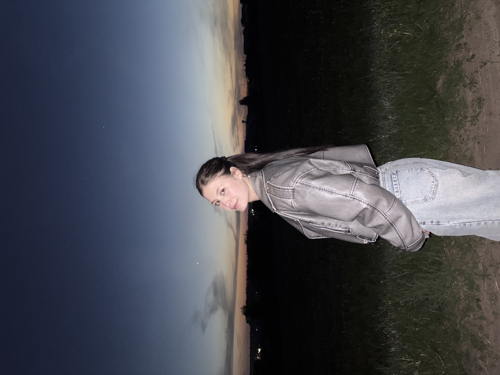 A young woman stands outdoors at dusk, wearing a gray jacket and light blue jeans. She is positioned with a landscape featuring the sky transitioning from dark blue to light orange during sunset. The lower portion of the scene consists of dark green vegetation and a dirt path. The image seems to have been taken facing the opposite direction of the subject, as it would normally appear.