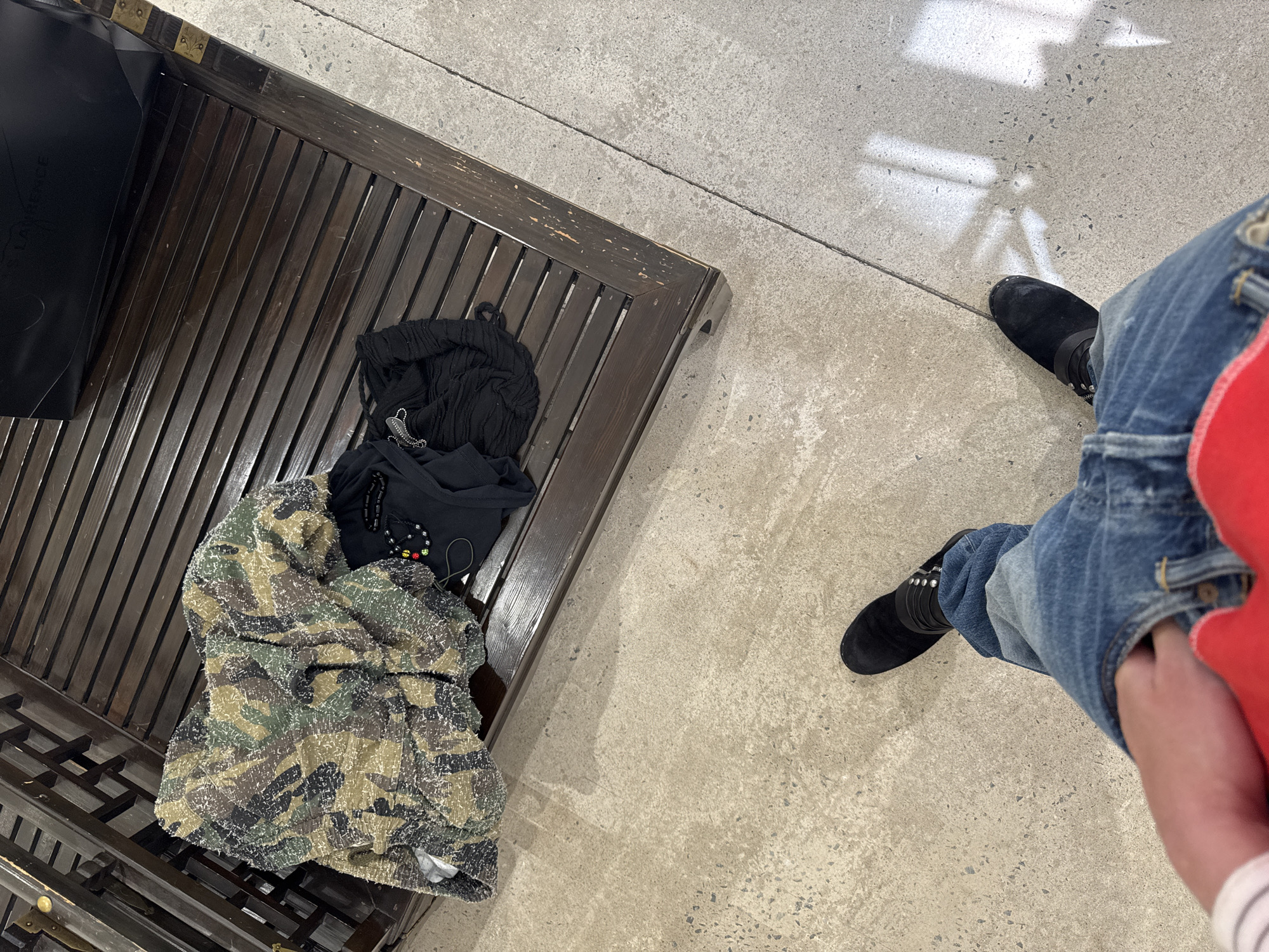 An overhead shot shows a portion of a slatted bench with discarded clothing and the lower portion of a person standing on a concrete floor. The bench is made of dark stained wood with decorative metallic accents, partially obscured by a black shopping bag and a pile of garments. The garments are comprised of a camo-patterned jacket or sweater, a black shirt with decorative stitching, and a black knitted hat or scarf. Adjacent to the bench, a person stands on the concrete floor, wearing blue jeans with rolled cuffs and black suede boots adorned with metallic studs. Their right hand is obscured inside the pocket of their jeans, and part of a red shirt or sweater is visible on their right side. The concrete floor extends across the frame, showing marks and patches consistent with a well-trafficked public space. No evidence indicates the city of Paris, France.