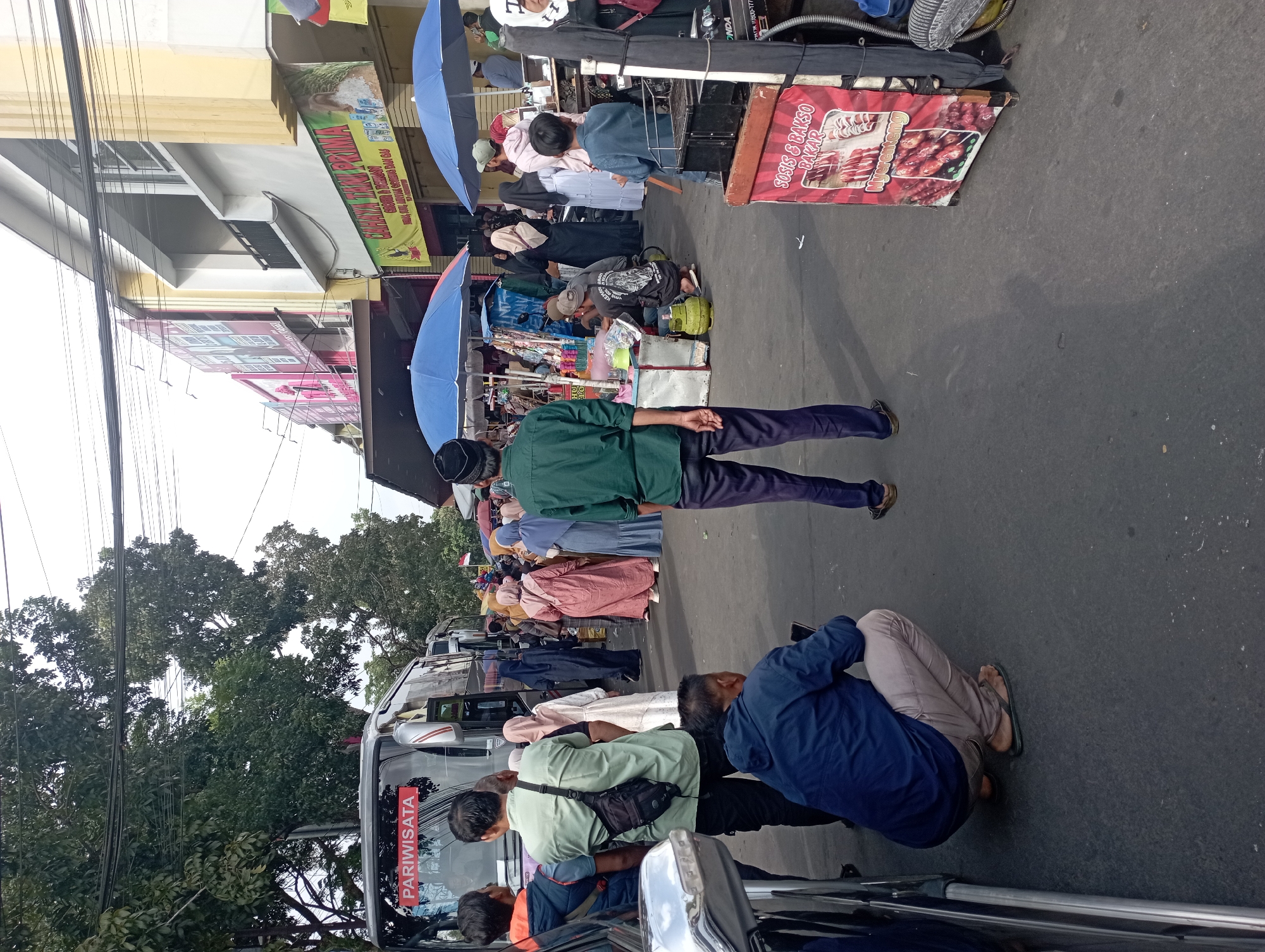 The image depicts a bustling street scene in Tasikmalaya, Indonesia, likely near a bus station or a market area given the presence of a "PARIWISATA" (tourism) bus and various vendors. The foreground shows several individuals, some standing and some squatting, observing the activity. A person in a dark blue jacket is squatting on the asphalt, looking at their phone. Further back, a man in a green shirt with a black sling bag stands facing away from the camera, and another man in a dark green shirt and pants is also facing away, wearing a black cap.
In the mid-ground, a group of people, predominantly women in hijabs and long dresses in various colors including pink, white, and blue, are gathered, possibly browsing or waiting. Two large blue umbrellas are set up, suggesting vendor stalls. One stall displays items for sale, and next to it, a green gas cylinder is visible, indicating cooking activity. Another vendor's cart is present, featuring a red banner with Indonesian text that appears to advertise grilled sausages and meatballs. The background reveals more buildings, including shops with colorful signage, and more people moving about. Overhead, a network of electrical wires crisscrosses the sky, typical of urban Indonesian settings. The overall atmosphere suggests a lively, public commercial area.