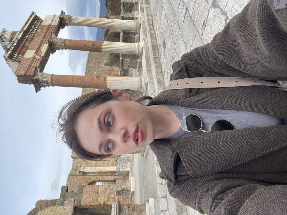 A young adult female is depicted in a mid-shot, slightly off-center to the left, positioned within the ancient ruins of Pompei, Italy. The specific location is the Forum, with the Temple of Jupiter partially visible in the background.
The female has fair skin, dark brown hair with loose strands, and light-colored eyes. Her gaze is directed upwards and to her left (viewer's right), not engaging directly with the camera. She is wearing a dark brown, textured double-breasted coat over a light grey ribbed top. A pair of aviator-style sunglasses with gold frames and dark lenses is hanging from her neckline. A light beige shoulder bag strap with dark circular perforations is visible across her right shoulder.
In the background, to the right, are three prominent fluted columns with Corinthian capitals supporting a partially collapsed architrave structure, constructed from a mix of brick and stone, characteristic of the Temple of Jupiter. The columns show sections of both white stone and reddish brick. To the left and further back, extensive ruins of Roman brick and stone walls with various openings are visible. The ground is paved with irregularly shaped, light-colored stone blocks. The sky is overcast and light grey-blue. Distant mountains or hills are discernible on the horizon, behind the ruins.
