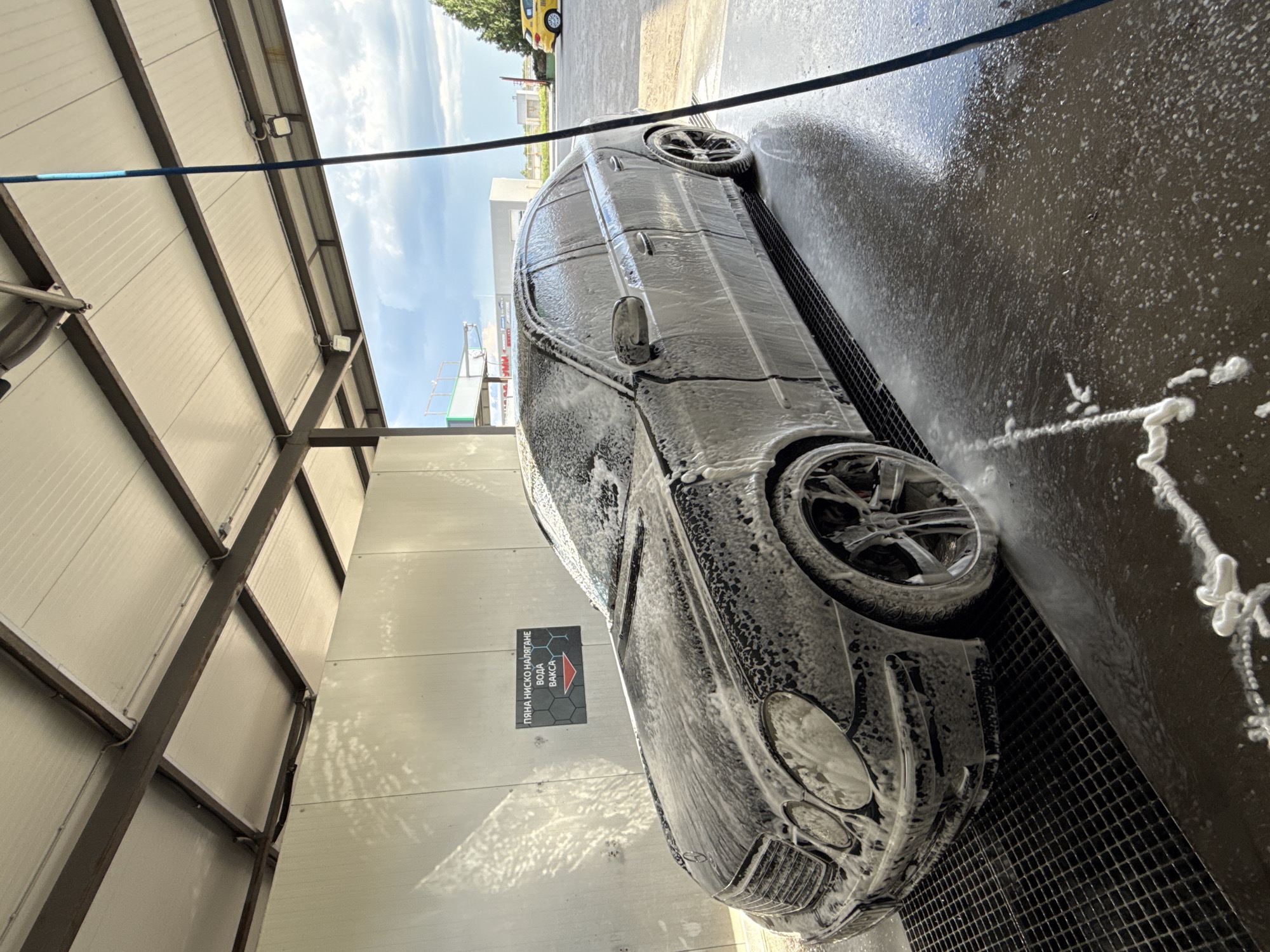 The image depicts a black Mercedes-Benz car being washed at a car wash. The car is covered in soap suds. The car wash facility has a corrugated metal roof and walls. A sign in Bulgarian is visible on the wall. The location is Stara Zagora, Bulgaria.