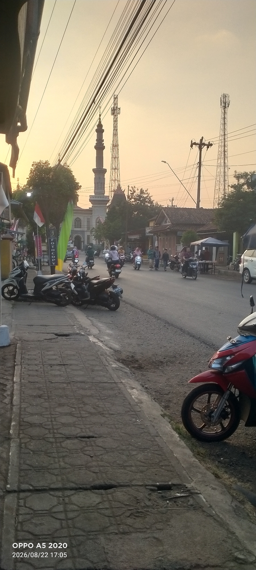 The image depicts a street scene in Kedung, Indonesia, with a mosque and communication towers visible in the background. The foreground shows a sidewalk with several parked motorcycles, and the street is busy with a number of people riding motorcycles. The sky is overcast with a warm, golden hue, suggesting it is either sunrise or sunset. The overall impression is of a typical street in a bustling Indonesian town.