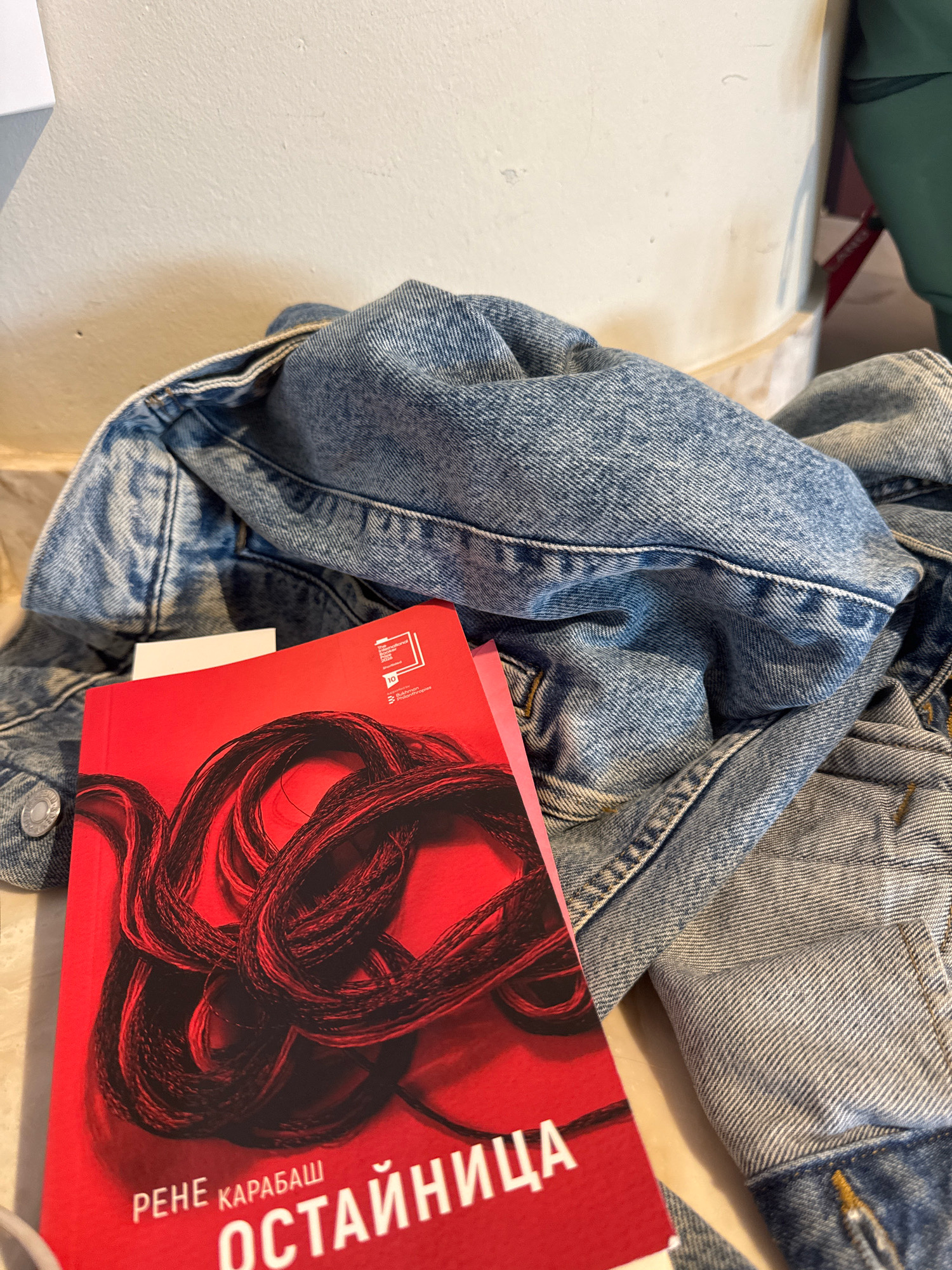 A close-up, eye-level shot shows a red book with a tangled, black cord design on its cover lying on top of a denim jacket. The jacket is partially unfolded and draped over a surface that appears to be a floor or low table, possibly in a room. The background is a neutral-colored wall, possibly concrete, with some subtle shadows. There are no people visible in the image. The book has Cyrillic text on it.