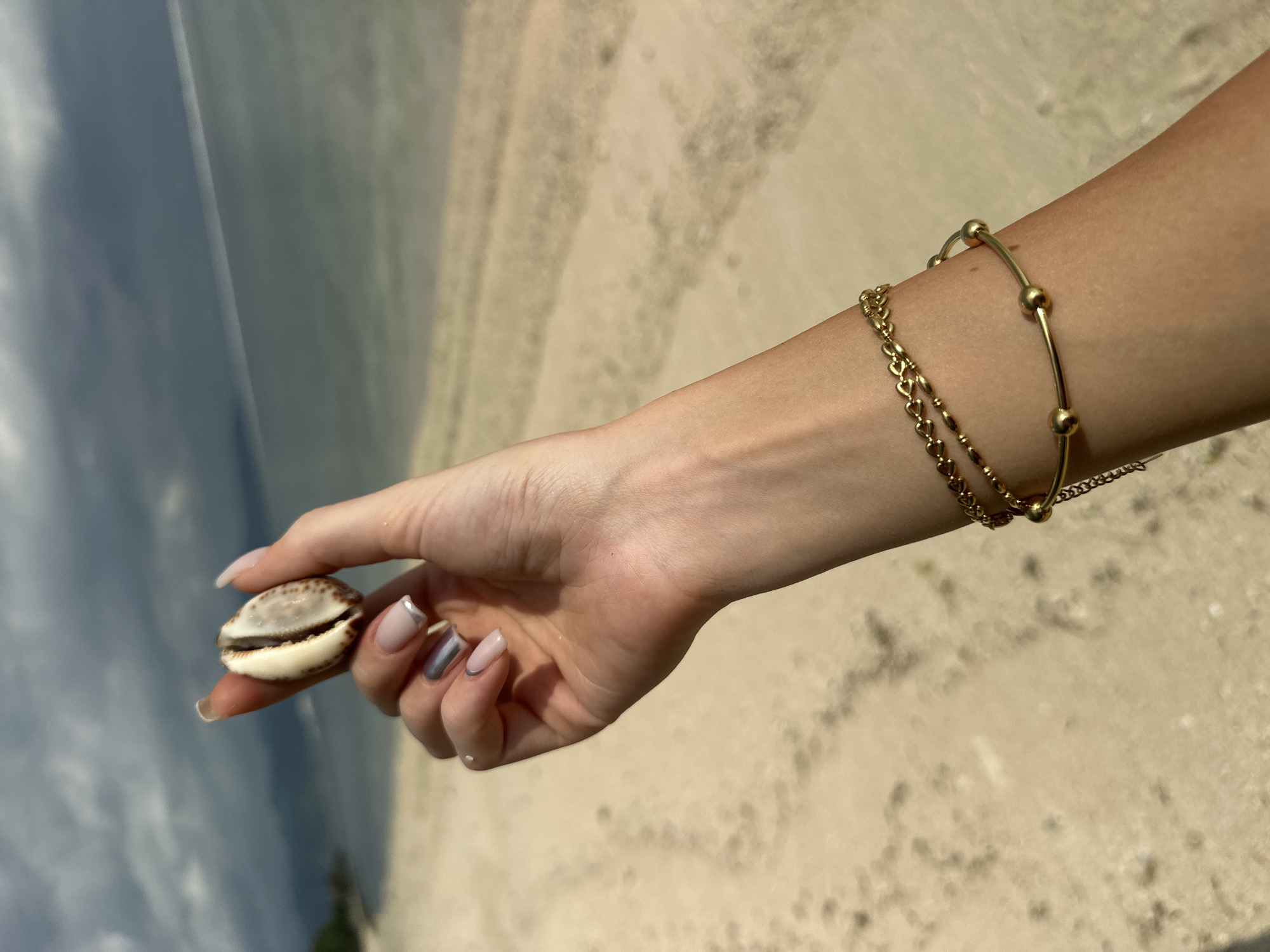 A person's right arm and hand are extended towards the left side of the image. The hand holds a cowrie shell between the thumb and index finger. The person wears two gold bracelets on their wrist. The fingernails have a light pink base color with silver tips. The background appears to be a beach with sand and water. The location is likely a beach in Unknown, Indonesia.