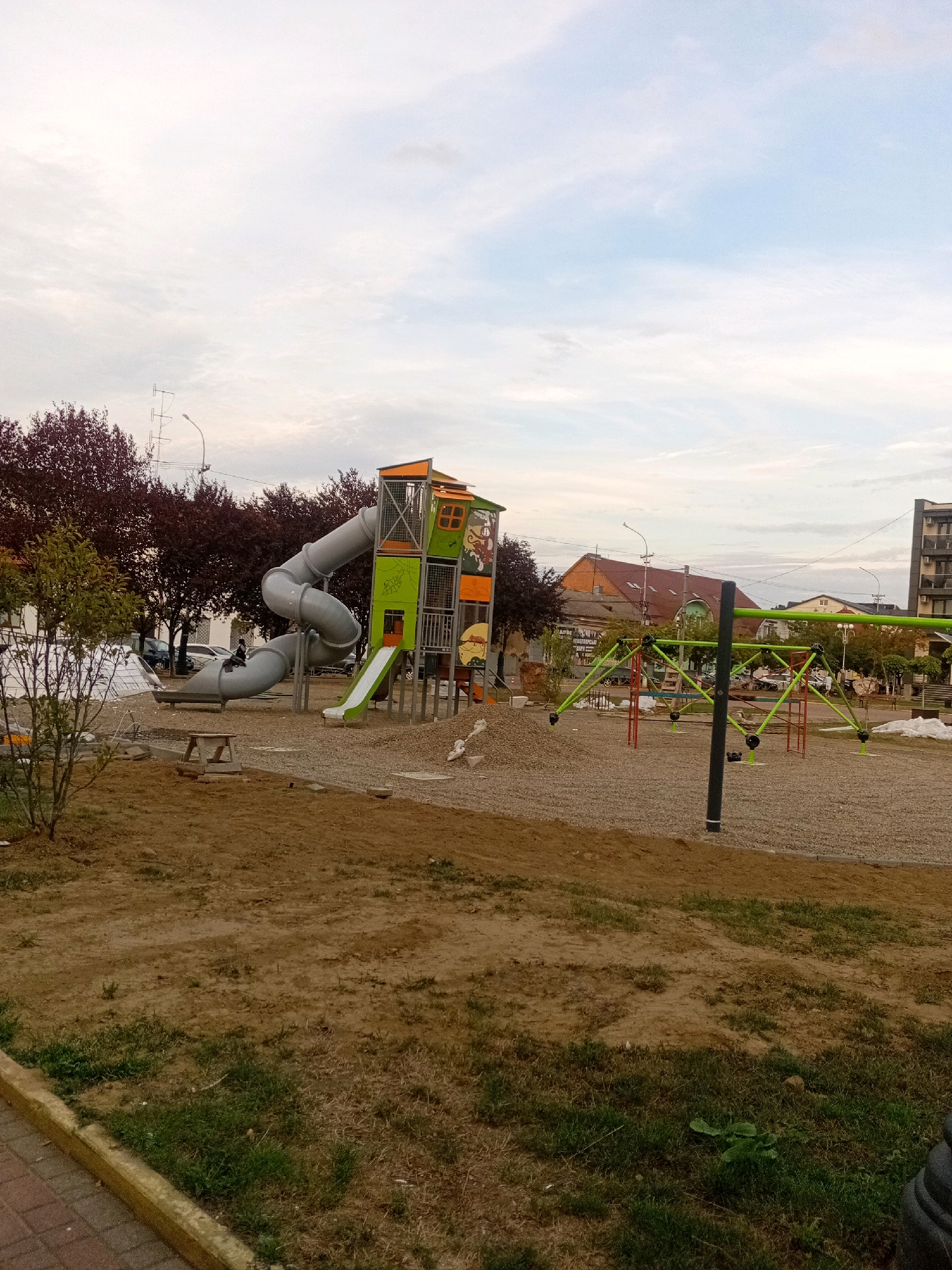The image depicts an outdoor playground, likely located in the city of Tiachiv, Ukraine, based on context not visible in the image itself. The playground features a large, grey, spiral tube slide connected to a colorful play structure. The structure has green and orange accents and appears to be multi-level, with various platforms and openings. Adjacent to the main structure, a smaller green slide is visible. In the foreground, there is an area of loose brown soil and scattered green grass. In the middle ground, a green geometric climbing frame with red supports is present, surrounded by gravel. In the background, there are trees with dark red foliage, buildings, and a cloudy sky. A single person is visible on the spiral slide. The playground appears to be under construction or recently completed, with some areas of exposed earth and loose gravel.