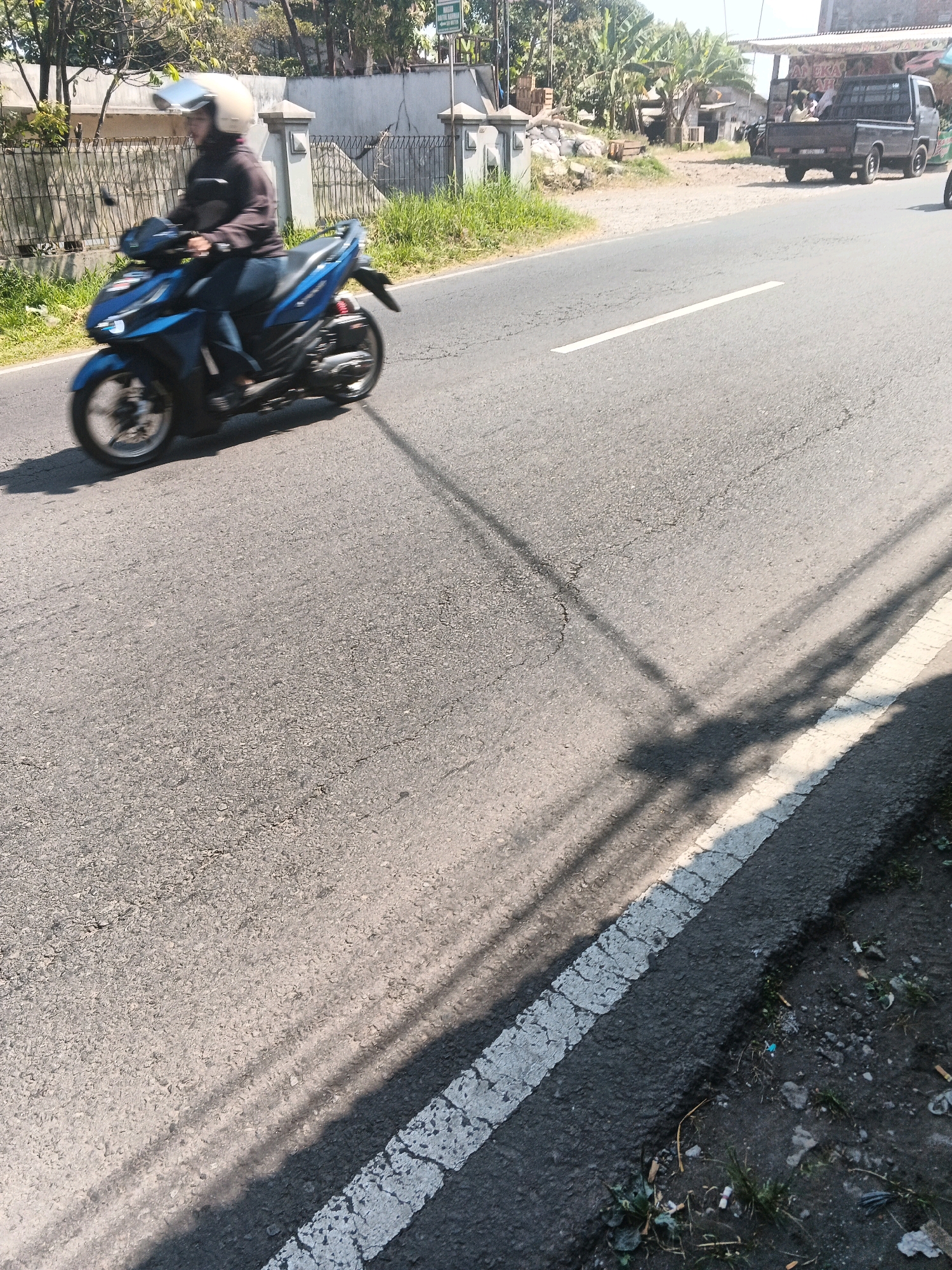 In Bandungan, Indonesia, on a sunny day, a person rides a blue motorcycle down a paved road. The rider wears a white helmet and dark clothing. The motorcycle has motion blur, indicating it is in motion. The road has a white line marking the lane. To the left of the road is a metal fence and vegetation. In the background, a black pickup truck is parked, and there are buildings and trees. The right side of the road has a dirt shoulder with some grass and debris. The lighting suggests it is daytime.
