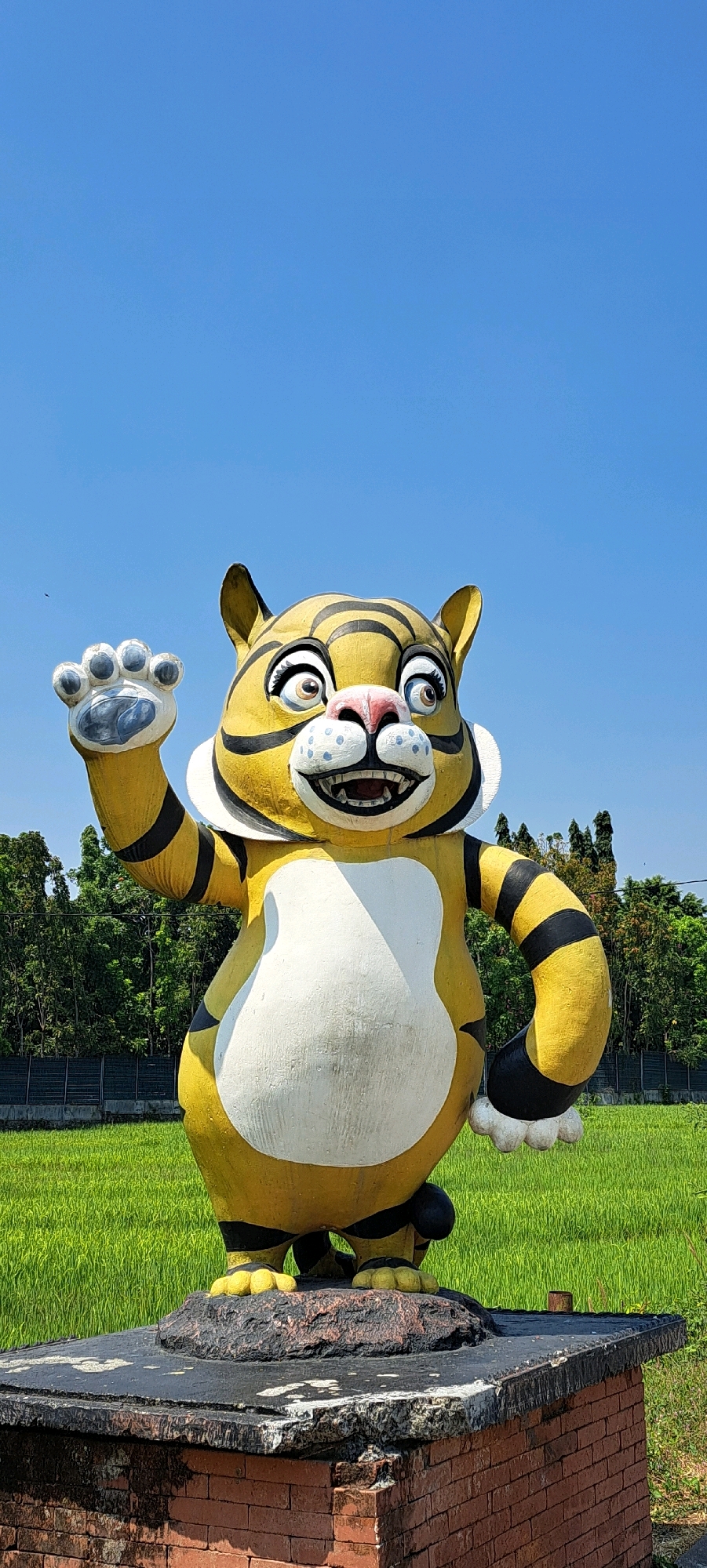 A statue of a cartoon tiger stands on a brick pedestal in a grassy field under a clear blue sky. The tiger is yellow with black stripes and a large white belly. It has a wide, smiling mouth, big eyes, and is raising its left paw as if waving. In the background, a line of green trees is visible, bordering the field. The pedestal is constructed from red bricks and has a weathered, dark gray top. The scene appears to be outdoors, possibly in a park or rural area. The location is identified as Lemahbang, Indonesia.