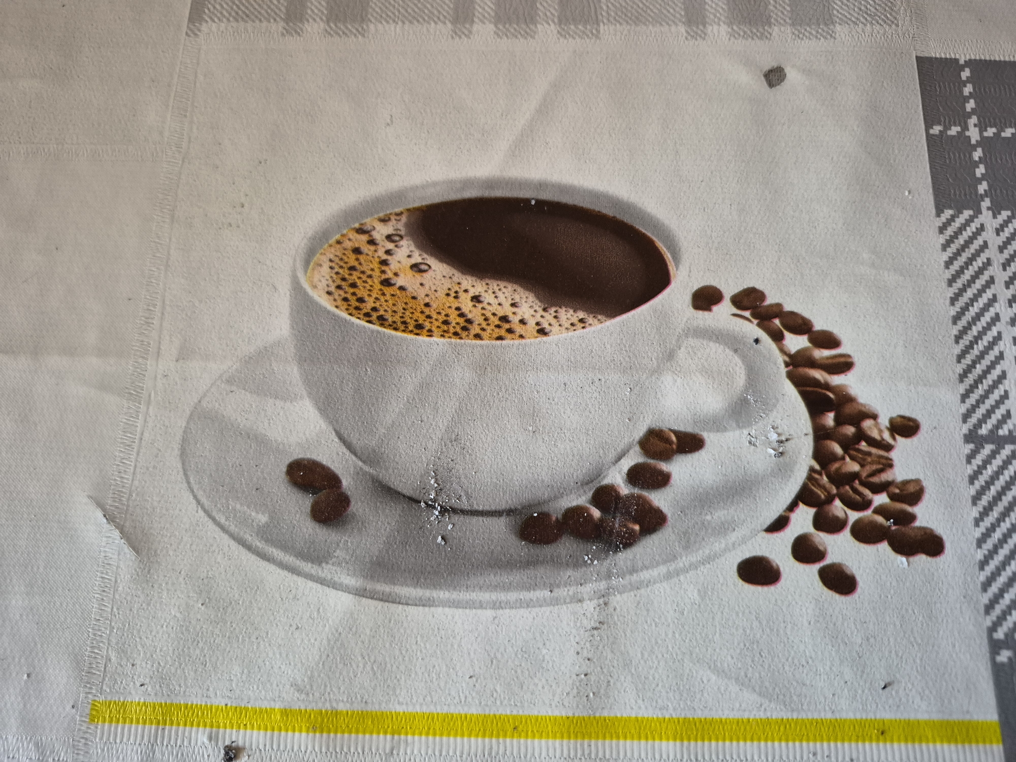 The image displays a close-up of a surface, likely a table mat or placemat, featuring a printed design of a coffee cup and coffee beans. The central subject is a white ceramic coffee cup, depicted from a slightly elevated angle, filled with dark brown coffee topped with a layer of lighter, frothy crema. The cup rests on a matching white saucer.
Scattered around the cup and on the saucer are numerous dark brown coffee beans, some individual and others clustered, extending towards the right side of the frame. The background surface is light-colored, appearing off-white or light grey, with a visible fabric-like texture. Subtle linear patterns are visible on the left side, resembling stitching, and a more distinct grey and white checkered pattern is present in the top-right corner.
A prominent horizontal yellow stripe runs across the bottom of the image, also exhibiting a textured edge, possibly indicating another seam or border. Small flecks and specks, which appear to be dust or debris, are visible on the surface of the mat, particularly near the cup and on the yellow stripe, suggesting this is a photograph of a physical object rather than a purely digital graphic.
No people are visible, and no specific activity is taking place beyond the static depiction of the coffee scene. There are no direct location cues within the image itself, and no visible text. The overall impression is a casual, inviting depiction of coffee, printed on a textured material.