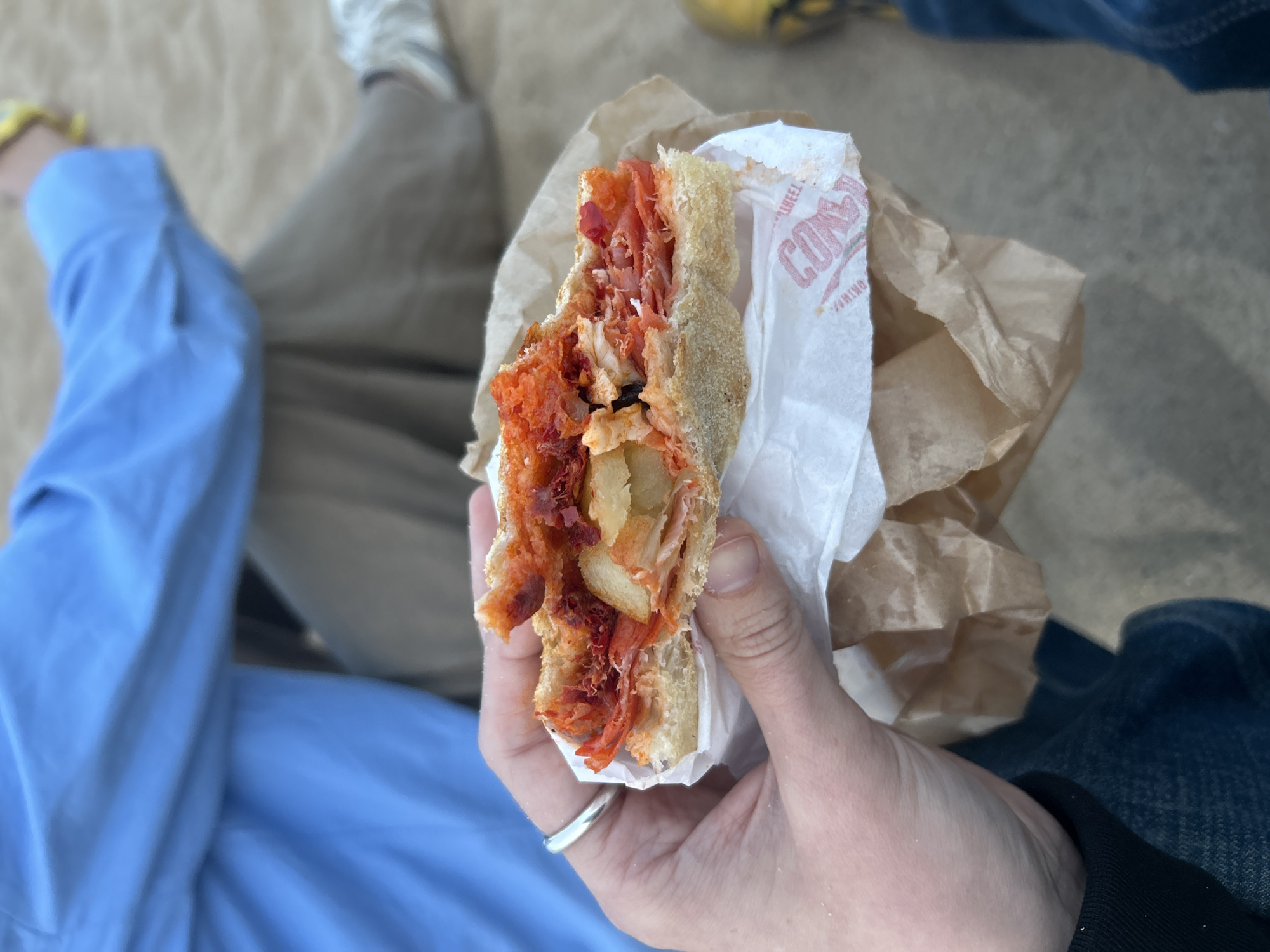 A person sitting on a sandy surface is holding a half-eaten sandwich in their hand. The sandwich is in a wrapper with the logo "CON" on it. The sandwich appears to have meat, what looks like potatoes, and some red sauce. The person is wearing a ring on their left ring finger. To the left of the person's hand, the person is wearing a blue shirt and khaki pants. To the right of the person's hand, the person is wearing blue jeans. It is a sunny day. A Conchino sandwich is a popular item to eat on Barceloneta Beach in Barcelona, Spain. The person holding the sandwich is sitting on the sand, likely on Barceloneta Beach.