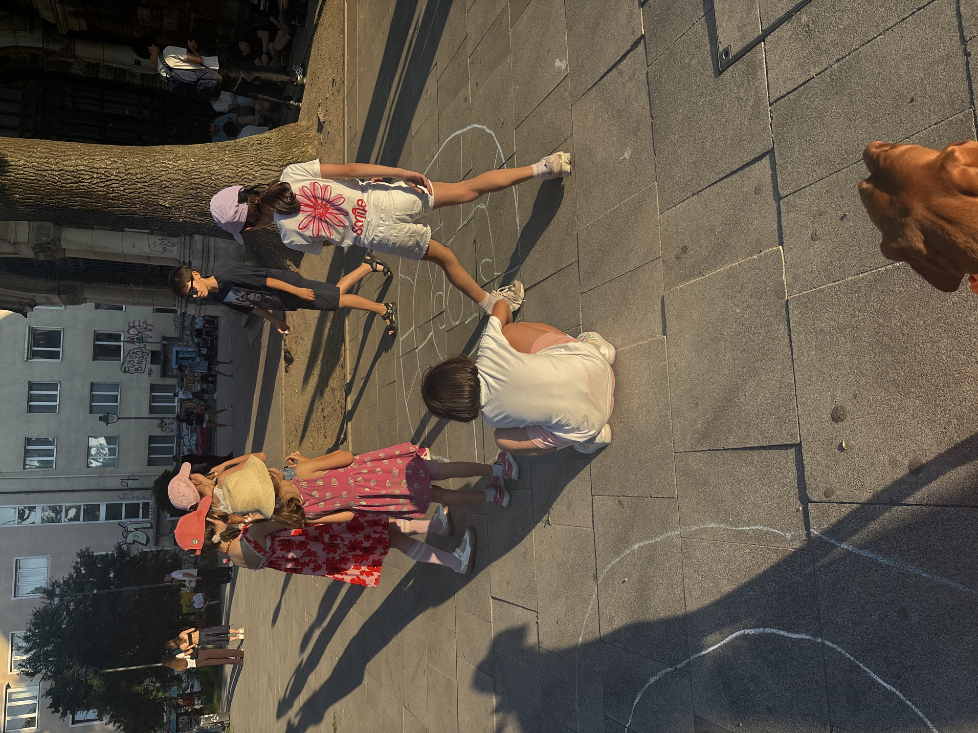A group of children are playing hopscotch on a paved city square in Sofia, Bulgaria, near a large tree and building facades. The children are scattered, with one child sitting and facing away from the camera, another standing with their back to the camera, and two girls in dresses and hats facing away. A boy in a dark t-shirt and shorts walks past in the background. The ground is marked with chalk lines for the game. The sun casts long shadows across the square, indicating it is late afternoon or early evening.