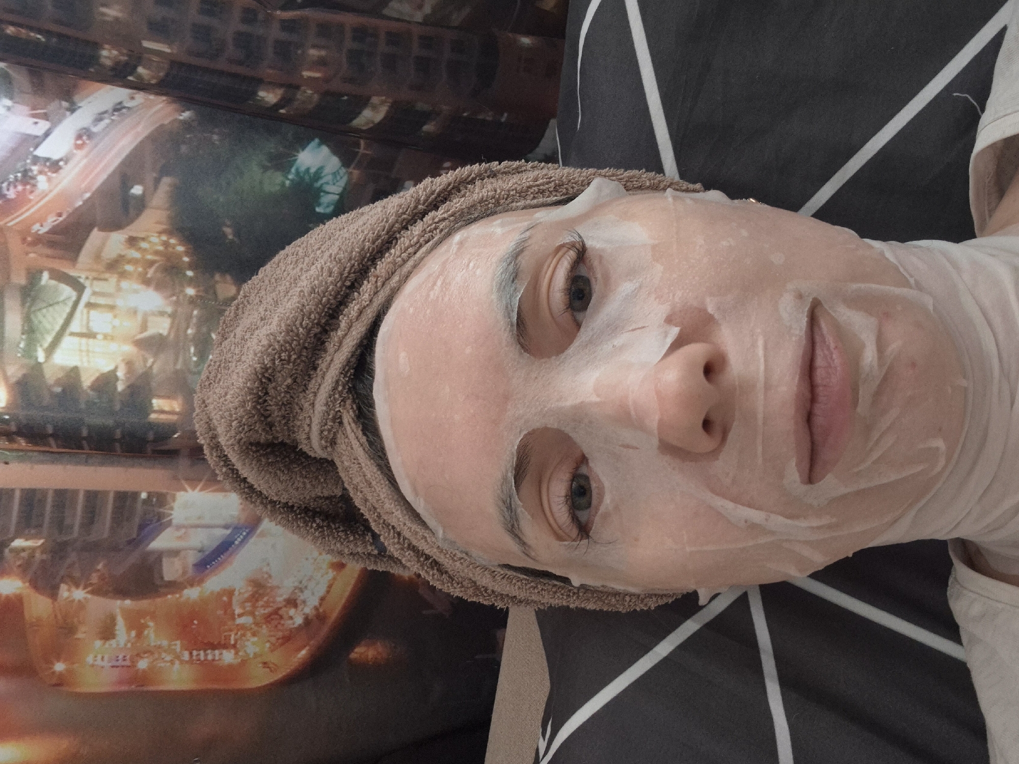 A person is lying on a bed, wearing a facial mask and a brown towel wrapped around their head. The bed is covered with a black and grey geometric pattern. The person's face is oriented towards the camera, and the camera angle is a high-angle shot. To the left of the person is a picture depicting a cityscape. Based on the location, the cityscape likely features elements of Chaikovsky, Russia. The person appears to be relaxing or undergoing a skincare routine.