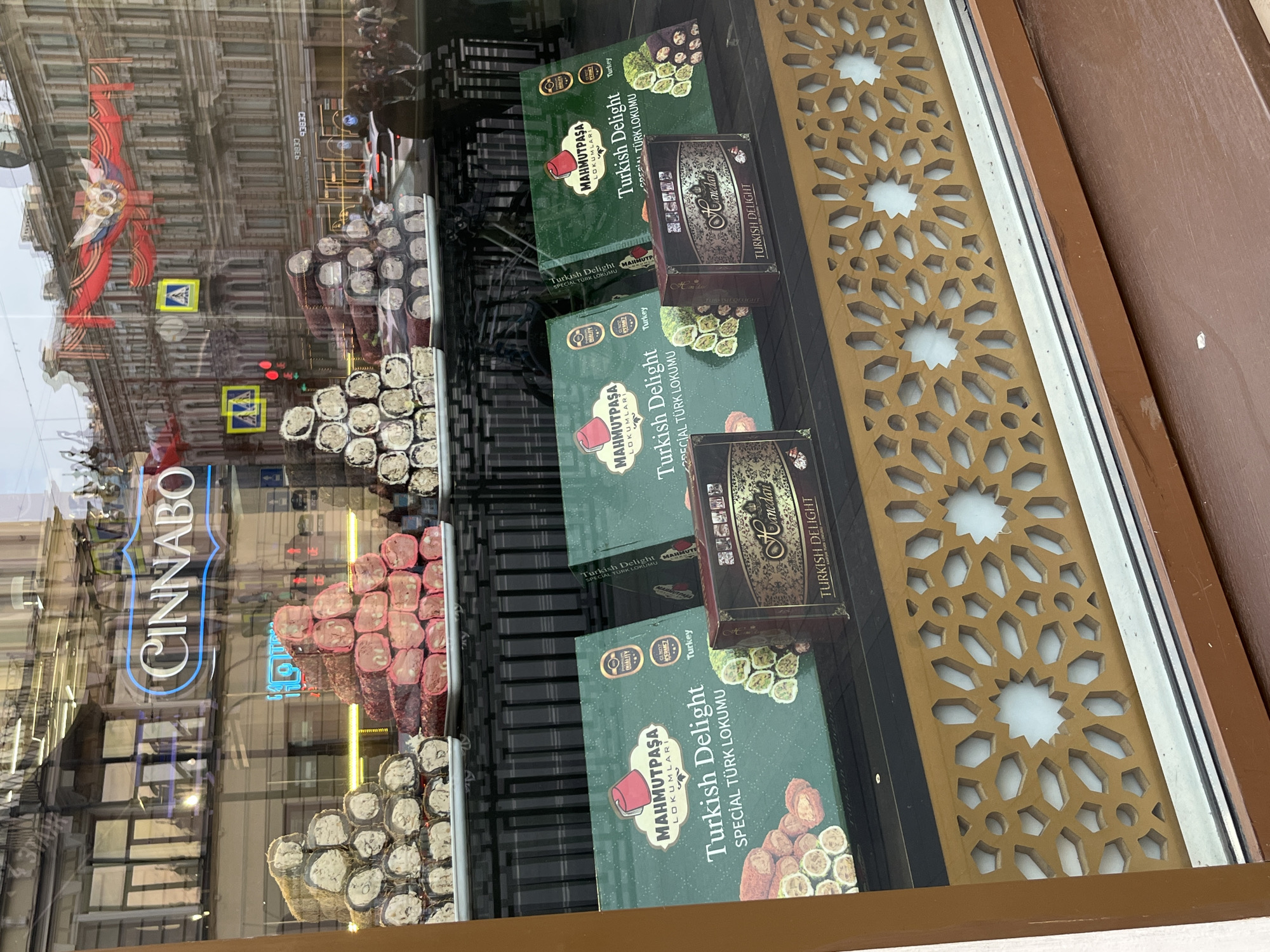 Here is a detailed description of the image:
**Scene:** The image is taken from within a shop or store, looking out through a window at a street scene. The shop's window display features various stacks and arrangements of Turkish Delight candies and boxes. The outer window reflects the urban landscape of a city.
**Subjects and Objects:**
* **Turkish Delight**: The main focus is on the Turkish Delight candies, arranged in rolls and piles.
* **Candy Boxes**: Boxes of Turkish Delight with the brand "MAHMUTPAŞA LOKUMLARI" are visible. These boxes feature text in Turkish and English, advertising "Turkish Delight" as "SPECIAL TÜRK LOKUMU". Some boxes include the word "Hanedan" as well.
* **Shop Window:** The window reflects the street outside, including buildings, traffic lights, street signs, and pedestrians. One building visible in reflection has a large red banner, and there is a store sign, partially obscured but readable as "CINNABO."
* **Store Interior:** Elements of the store's interior are visible, including shelves and decorative elements like an ornate gold-colored cutout design near the window edge.
**Actions and Interactions:**
* The photo captures a static scene of a shop display, with an implied action of a customer potentially viewing the products from the street.
**Environment:**
* **City:** The reflected buildings and traffic indicate a busy urban setting.
* **Shop/Store**: The context is a retail environment, presumably a candy store or a shop selling Turkish delicacies.
* **Time of day**: Judging from the light, it appears to be daylight.
**Location:**
* Based on the product branding and street views, it is likely that this scene is in Turkey.
* The Cinnabo sign in the window may refer to a restaurant chain.
Overall, the image presents a shop selling Turkish Delight, captured through the window with a background of a bustling city street.