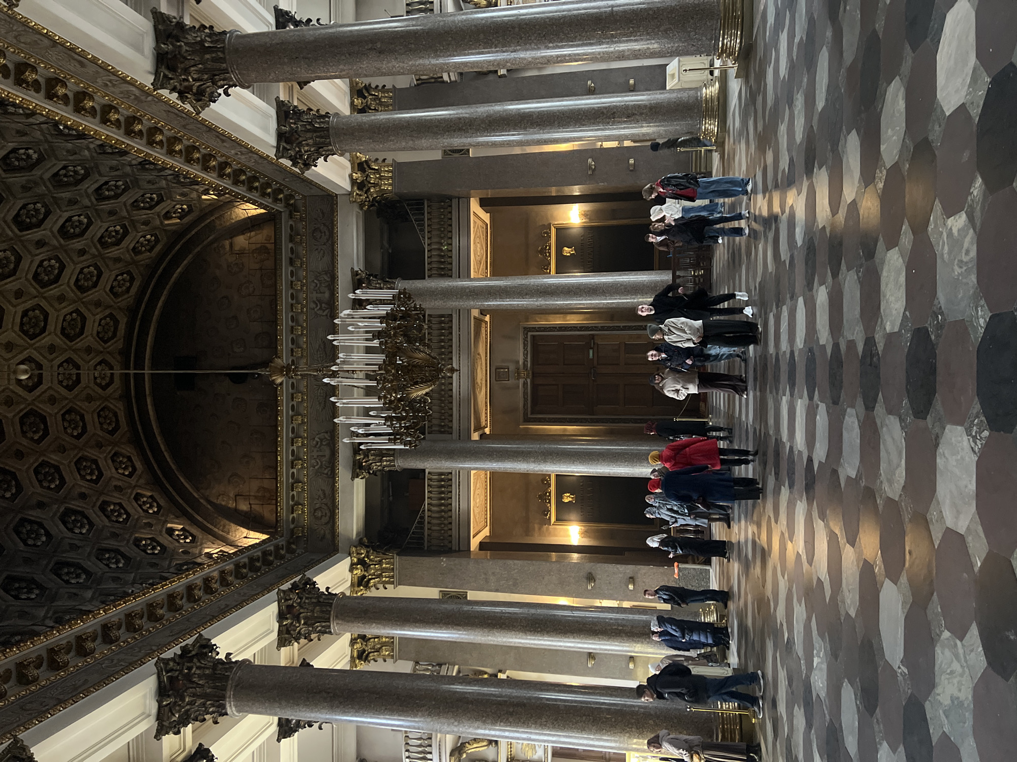 The image shows the interior of Saint Isaac's Cathedral in Saint Petersburg, Russia. A group of people, likely tourists, stand in the central nave, facing away from the camera. They appear to be listening to a guide. The architecture is grand, with tall marble columns topped with ornate capitals that line the nave, leading the eye towards a vaulted ceiling and dome. A large chandelier hangs from the ceiling. The floor is patterned with tessellated stone tiles. There are doorways in the background with ornate frames.