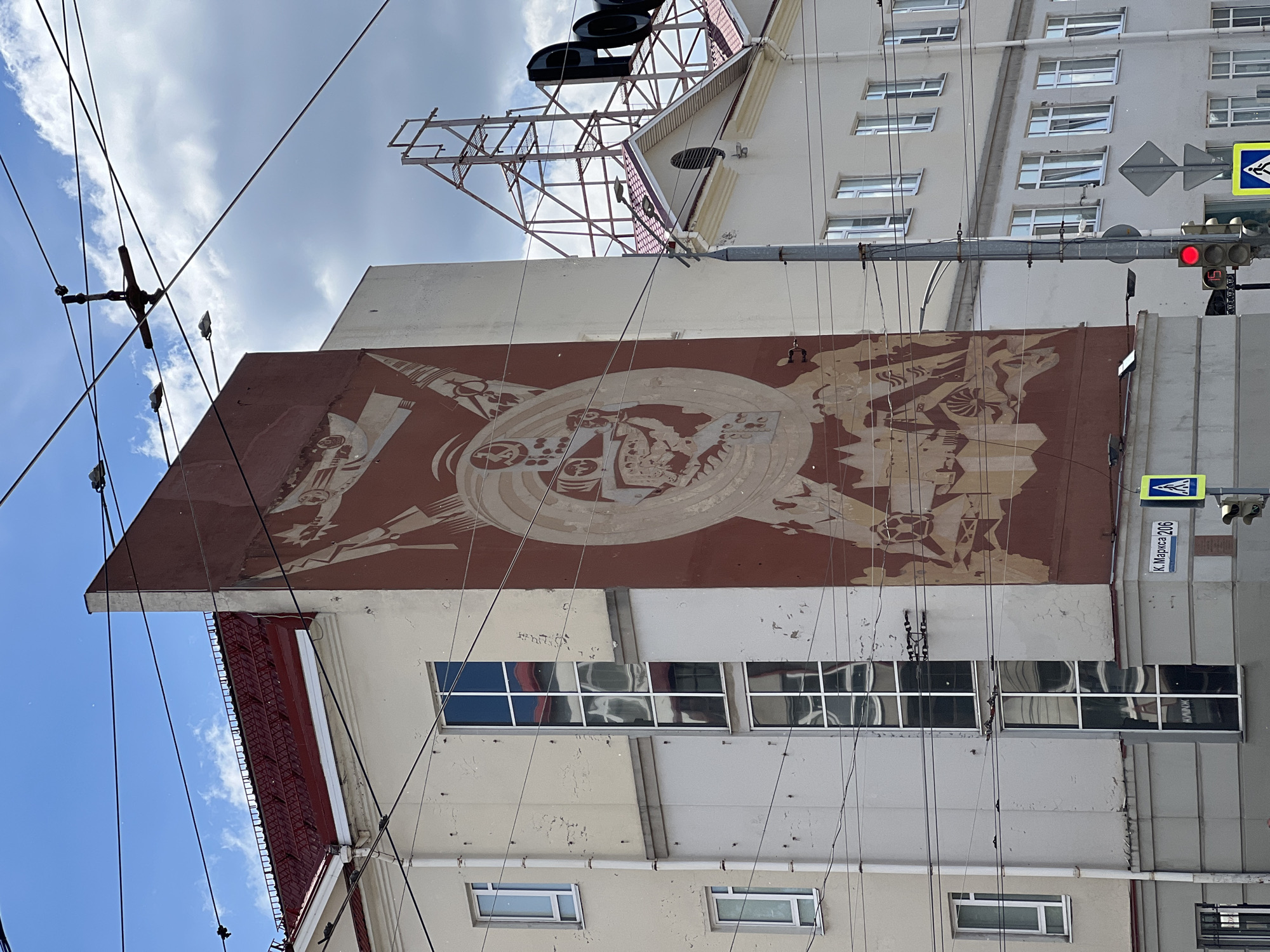 This upward-angled outdoor shot captures the facade of a multi-story building in an urban setting, likely Izhevsk, Russia. The building features a prominent, large-scale mural rendered in reddish-brown and cream tones, depicting stylized, possibly industrial or socialist realist, figures and symbols arranged in a circular motif. The mural shows signs of age, with some fading and wear.
Above the mural, the building's light-colored facade appears aged with peeling paint and staining. Multiple windows, some reflecting the blue sky and clouds, are visible. Below the mural, a reddish, ornate roofline or decorative element adds architectural detail. At the very top of the building, a large skeletal metal structure, possibly for a billboard or antenna, is visible, with large, black, letter-like shapes (perhaps "ДОБ" or similar) mounted on it.
Numerous black power and communication lines crisscross the upper part of the image, leading to poles and connecting to the building. The sky is partly cloudy with patches of bright blue. On the right side of the image, a traffic light displays a red signal. Below it, a blue street sign with white Cyrillic text reads "К. Маркса 206", indicating "K. Marx 206" (Karl Marx Street, building number 206). Above the street sign, a standard yellow and blue pedestrian crossing sign is visible. The scene is captured during daylight hours.