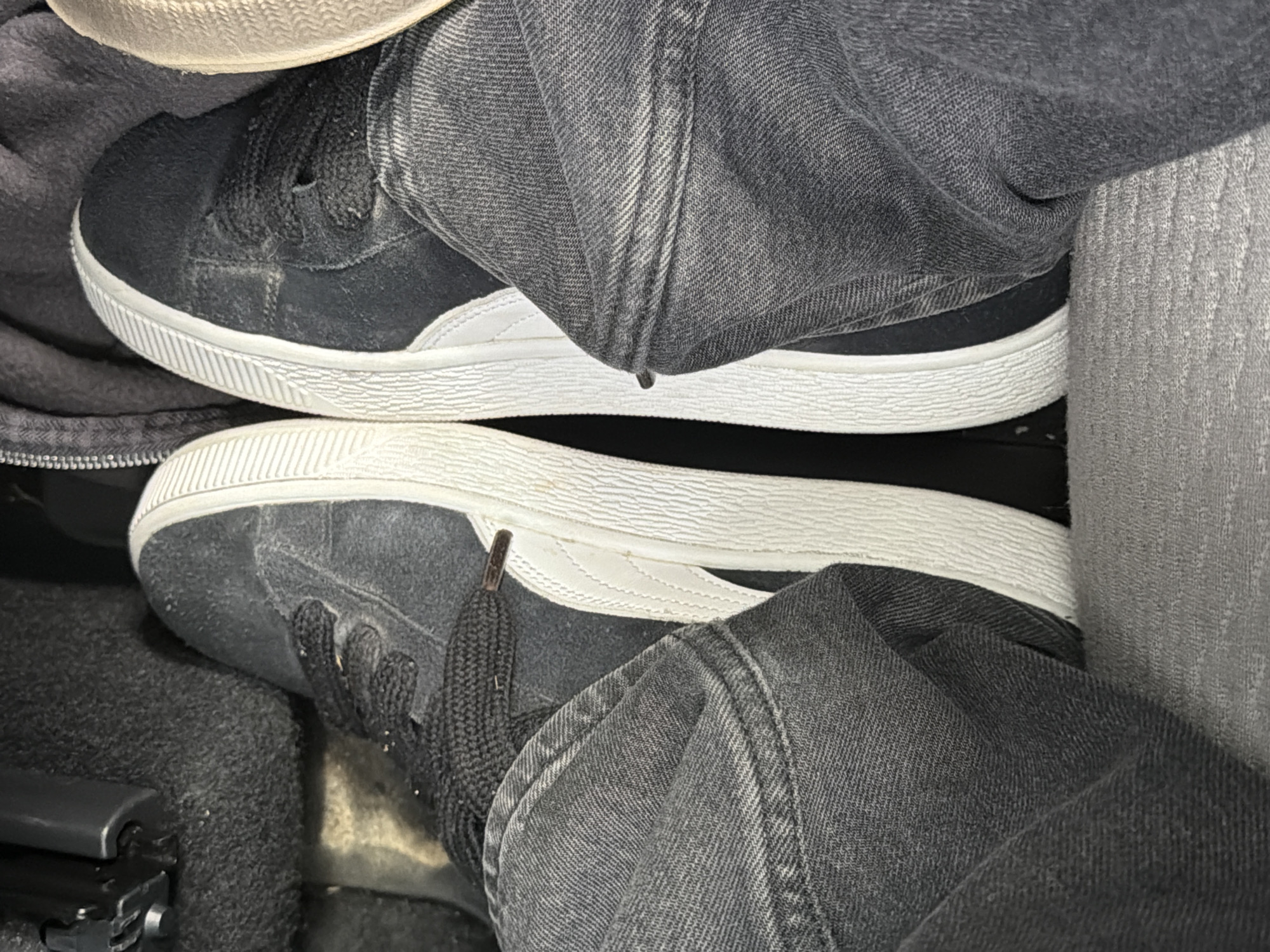 The image shows a close-up of a person's feet wearing black Puma suede sneakers and dark grey jeans. The shoes have white rubber soles and white accents on the sides. The person's feet are positioned as if they are in a car, with the floor mat and part of a car's interior visible. The perspective is from above, looking down at the feet. There is no indication of the specific location being Vallensbæk Landsby, Denmark.