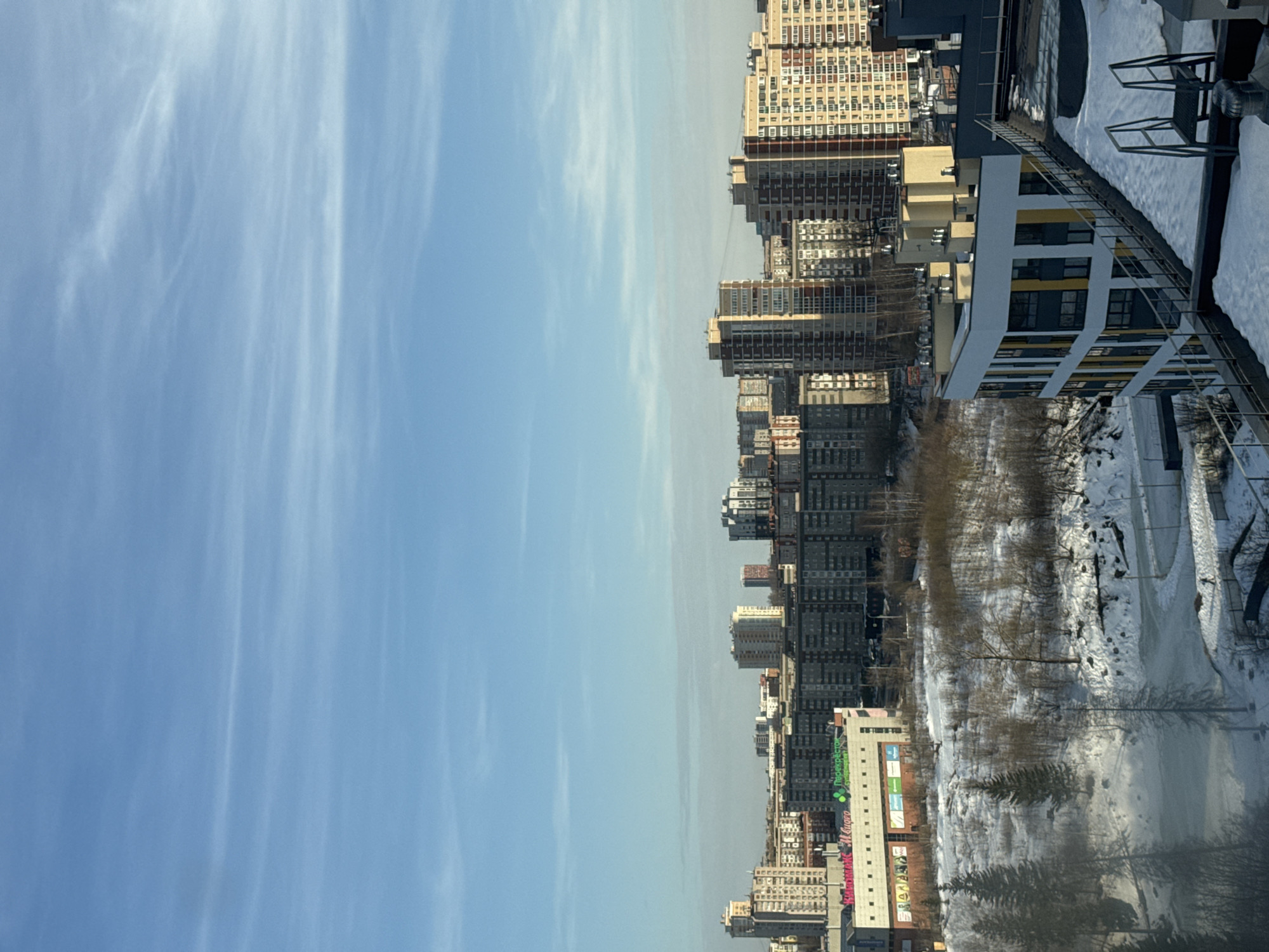 This image captures a wide-angle view of a winter cityscape in Izhevsk, Russia, under a clear blue sky with sparse, wispy cirrus clouds. The scene is illuminated by natural sunlight, indicating daytime.
In the immediate foreground, the snow-covered rooftop of a modern building with a distinctive curved facade is visible. This building features vertical sections of yellow, white, and dark grey paneling. A metal railing extends along the edge of the rooftop, and a dark metal ladder-like structure stands on the snow-covered roof surface in the lower right.
The mid-ground reveals a snow-covered, undulating landscape interspersed with deciduous trees, now bare, and several dark green coniferous trees. A winding, snow-laden path or road traverses this area, appearing to border a partially frozen or snow-covered body of water on the left. A single, dark human figure is discernable walking along one of these snowy paths in the lower mid-ground.
Beyond this natural expanse, a large, multi-story commercial building stands prominently. Its facade displays signage for "ПЕРЕКРЕСТОК" (Perekrestok, a supermarket chain) in green and "М.Видео" (M.Video, an electronics retailer) in red, indicating a shopping center.
The background is dominated by a dense cluster of multi-story urban residential buildings. These vary in height and architectural style, ranging from older, darker grey concrete structures to more modern buildings with lighter-colored facades, including some with prominent beige and reddish-brown sections. The city extends broadly under the bright winter sky.