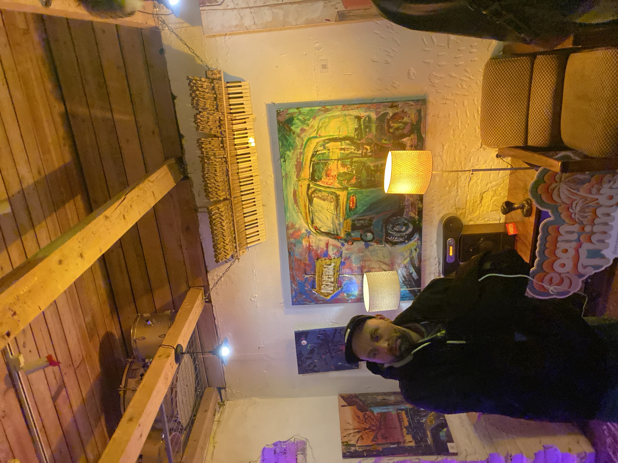 The original image is rotated 90 degrees clockwise. When viewed correctly, it depicts an interior scene, possibly a creative studio, bar, or cafe in Moscow, Russia, filled with rustic and artistic elements.
A man is positioned on the right side of the frame, captured from the chest up, looking directly at the camera with a neutral to slightly smiling expression. He has a dark beard and is wearing a dark cap and a black hooded jacket.
The space features a mix of exposed wooden plank walls and light-colored, textured brick or plaster walls. Several artworks adorn the walls, most notably a large, vibrant abstract painting in the center depicting a colorful auto-rickshaw or similar vehicle. Below it, two smaller abstract paintings are visible: one predominantly dark blue and another featuring earthy tones, possibly a landscape.
The room is illuminated by warm, artificial light. Two wall-mounted lamps with yellow, woven shades provide a soft glow, and a bare bulb hangs from an overhead wooden beam. A purple light source, possibly an LED strip, casts a hue on a lower section of the wall.
Distinct decorative elements include a unique wall hanging resembling multiple rows of miniature wooden piano keys on the left. In the upper left, parts of a drum kit (snare and kick drum) are visible. A beige, textured sofa or bench sits on the right. A skateboard leans against a wall, and a colorful cutout sign featuring partial text, "SOAN THIN" or "SOON THING," is also present. The scene suggests an evening or nighttime setting due to the reliance on artificial lighting, creating a cozy and relaxed artistic atmosphere. Some indistinct text, possibly "RIVEBC," can be seen on the large painting.