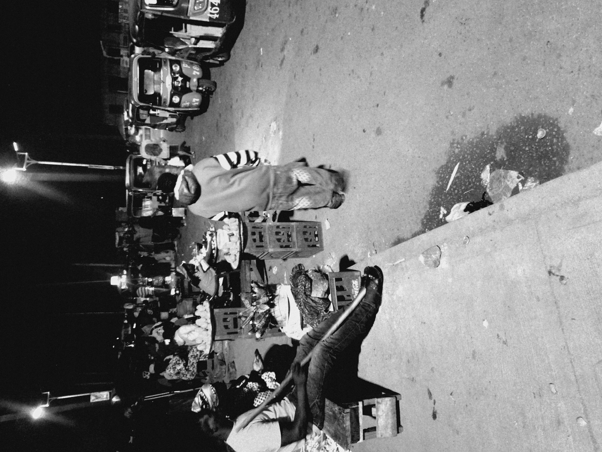 The image is a black and white photograph, possibly taken at night, depicting a street scene in Bukuru, Nigeria. Several auto-rickshaws are parked along the edge of the street. Adjacent to the vehicles, people are sleeping on top of stacked plastic crates and/or blankets. A street lamp illuminates the scene on the left side of the image. Trash and debris are visible on the street.