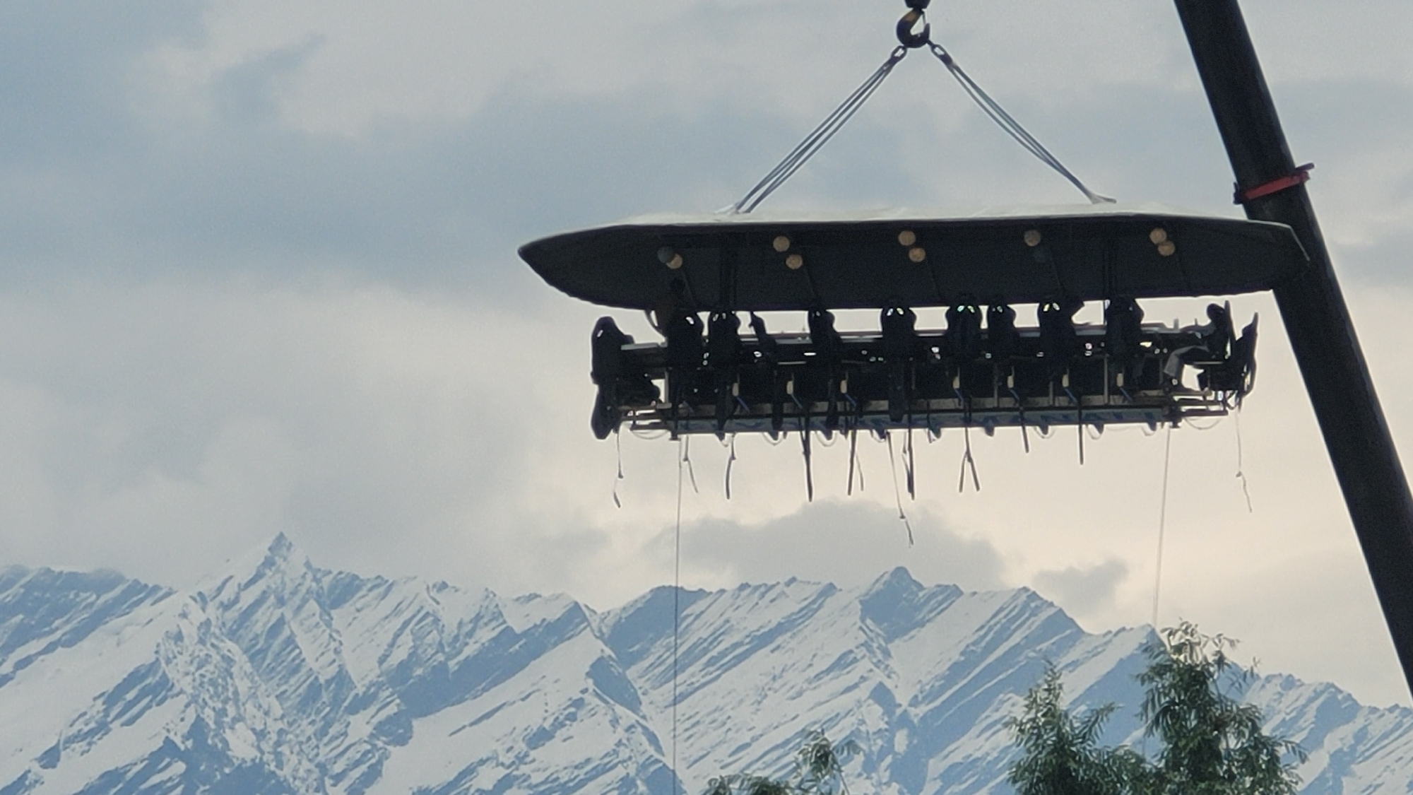 In Vashisht, India, a unique dining experience unfolds as a platform hosting diners is suspended in the air by a crane against a backdrop of snow-capped mountains and a cloudy sky. The platform, resembling an elongated table with chairs, is dark in color and appears to be supported by cables attached to a crane arm visible on the right side of the frame. Several people, silhouetted against the sky, are seated at the table, some appearing to be engaged in conversation or enjoying the meal. The mountains in the background are imposing, with sharp peaks and a covering of snow, suggesting a high-altitude location. The overall scene presents a surreal and adventurous dining setup amidst a dramatic natural landscape.