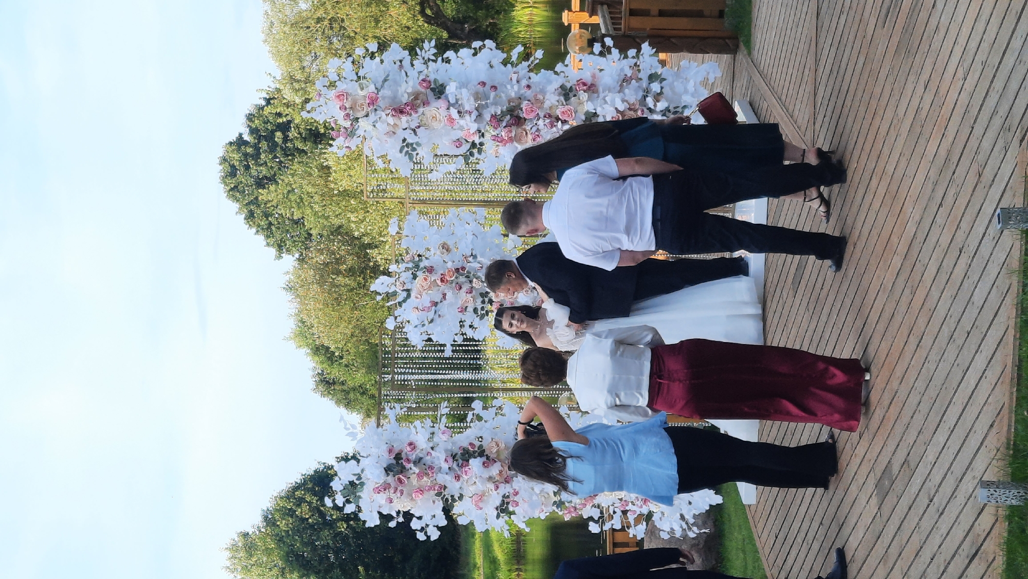 In Жодзішкаўскі сельскі Савет, Belarus, a wedding ceremony takes place outdoors on a wooden deck near a body of water. A wedding arch decorated with white and pink flowers serves as the backdrop for the couple. A bride in a white wedding dress and a groom in a dark suit stand in front of the arch. Several guests are present, with some observing the ceremony and others taking photos. A woman in a light blue top and black pants is capturing the moment with a camera. Another woman in a white top and a long, dark red skirt stands nearby. A man in a white shirt and black pants is positioned in front of the groom, facing away from the camera. To the right of the groom, a woman in a dark teal dress stands with a red purse. The surrounding environment includes lush green trees and a clear sky.