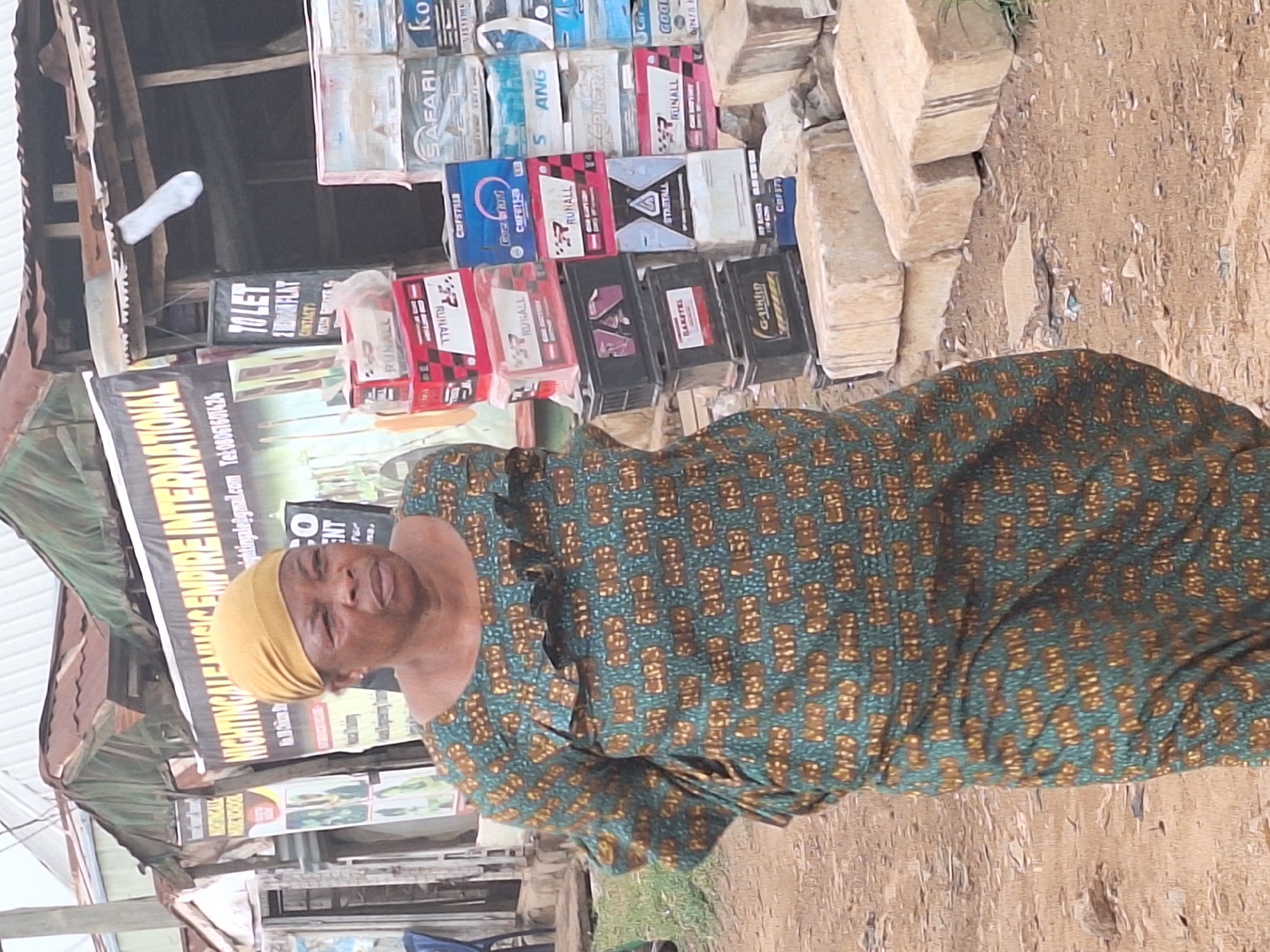 A woman with a yellow head covering and wearing a green and brown patterned dress stands outdoors in Sabon Tasha, Nigeria. Behind her are stacks of concrete blocks and boxes of car batteries. A sign for "NIGHTINGALE EMPIRE INTERNATIONAL" is visible, featuring contact information. The environment suggests a commercial or roadside sales area.