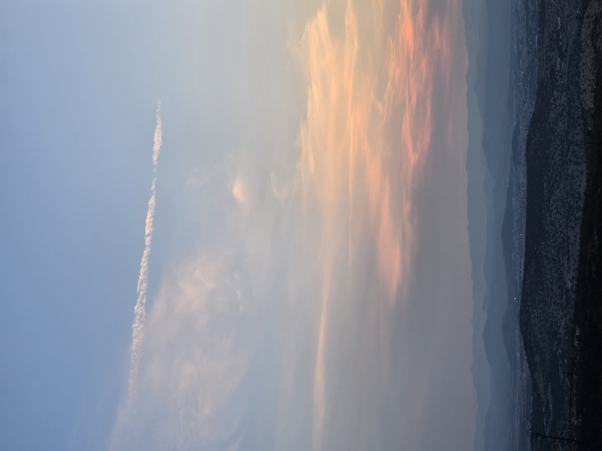 The image depicts a broad, panoramic view of a valley landscape at sunset. In the foreground, dark, undulating hills are covered with sparse vegetation, suggesting a dry, possibly arid environment. The middle ground features a sprawling urban area, with buildings and infrastructure faintly visible through the haze. Beyond the city, a series of layered mountain ranges recede into the distance, their forms softened by atmospheric perspective and the fading light. The sky above is a canvas of soft pastel colors, transitioning from a pale blue at the top to hues of pink and orange closer to the horizon, indicative of a sunset. A thin, elongated cloud stretches horizontally across the upper part of the sky. The scene is devoid of any visible human figures or specific actions. The overall impression is one of natural beauty and quietude, with the urban settlement integrated into the vast, mountainous terrain.