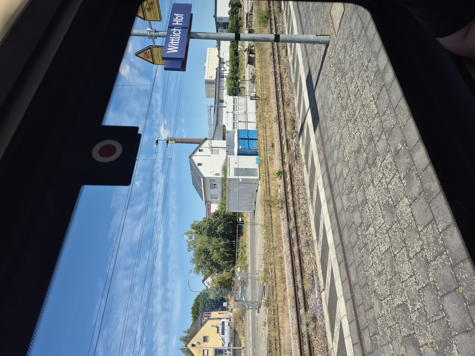 The image shows a view from inside a train looking out at a train station. A blue sign with white text reads "Wittlich Hbf", indicating the station's name. There are train tracks in the foreground, a paved platform, and beyond that, buildings and vegetation. Overhead, power lines are visible against a blue sky with scattered clouds. The scene appears to be in Germany.
