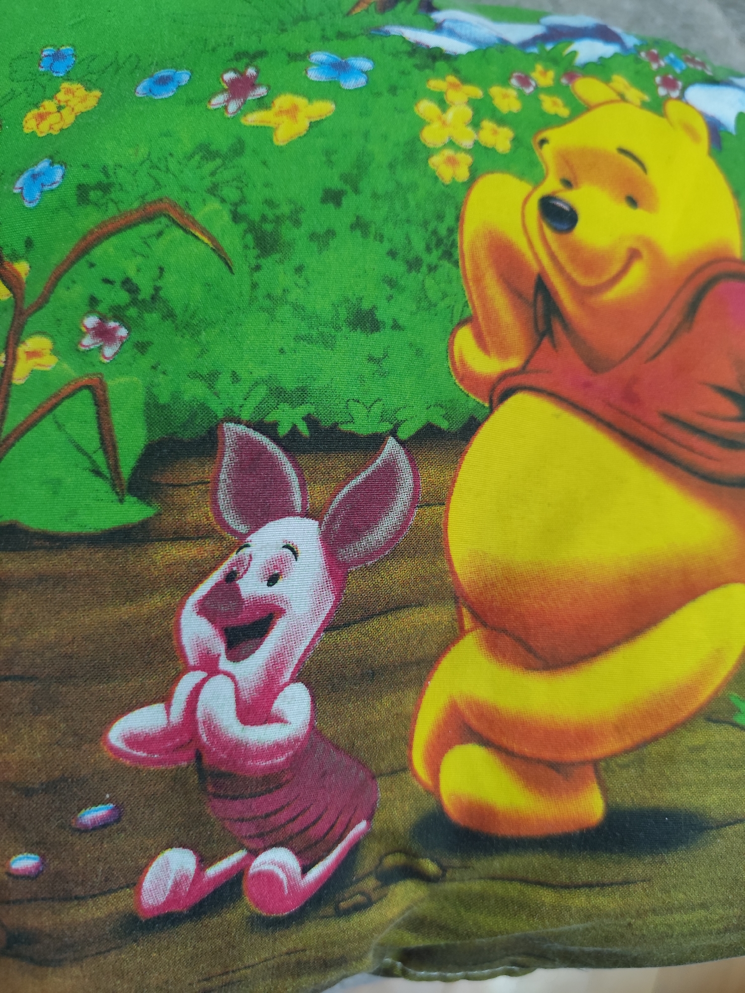 The image depicts a close-up of a children's fabric print featuring characters from Winnie the Pooh. In the foreground on the right, Winnie the Pooh is depicted with a smiling expression, leaning back and resting his arm behind his head. He is wearing his signature red shirt. To the left of Winnie the Pooh, Piglet is shown with his hands clasped together, looking happy and excited. The background is a green grassy area with stylized yellow and blue flowers, and a brown wooden surface that could represent the ground or logs. There are no elements in the image that indicate a specific location in Montana, Bulgaria, or any urban setting. The scene is a static illustration on fabric, not a real-world environment.