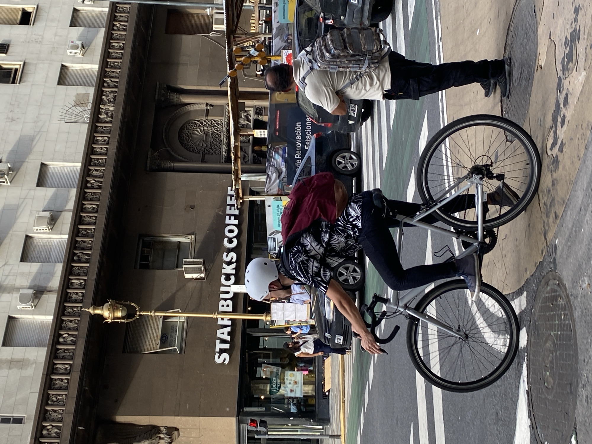 A person wearing a white helmet and a black and white shirt rides a gray bicycle on a street marked with white and green stripes. A Starbucks Coffee shop is visible in the background. A person wearing a backpack stands on the sidewalk in front of a dark-colored vehicle. The building facade beside the Starbucks has intricate architectural details and multiple windows. The image appears to be taken in Buenos Aires, Argentina, given the architectural style and the Spanish language signage.